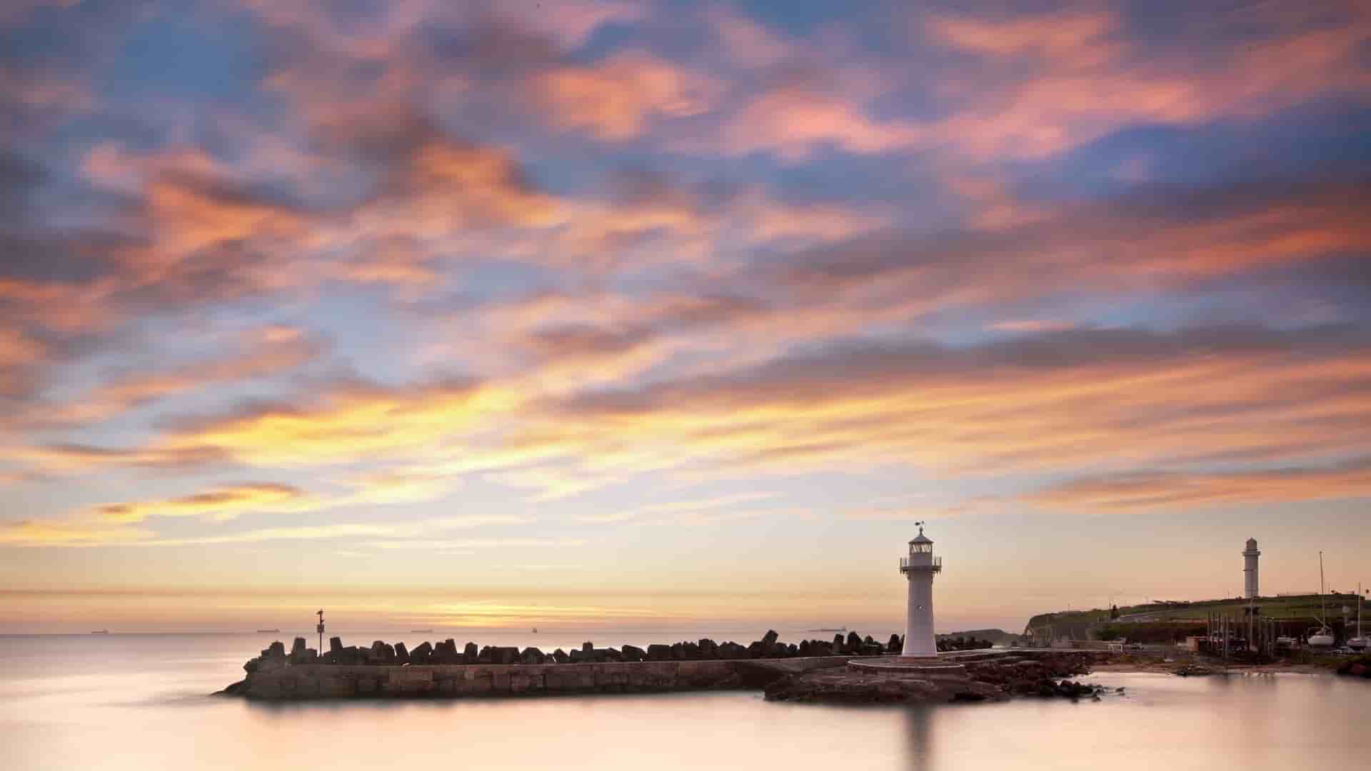 A stunning sunrise over a tranquil sea, with a lone lighthouse standing on a rock breakwater, with orange and yellow clouds on Norfolk Island.