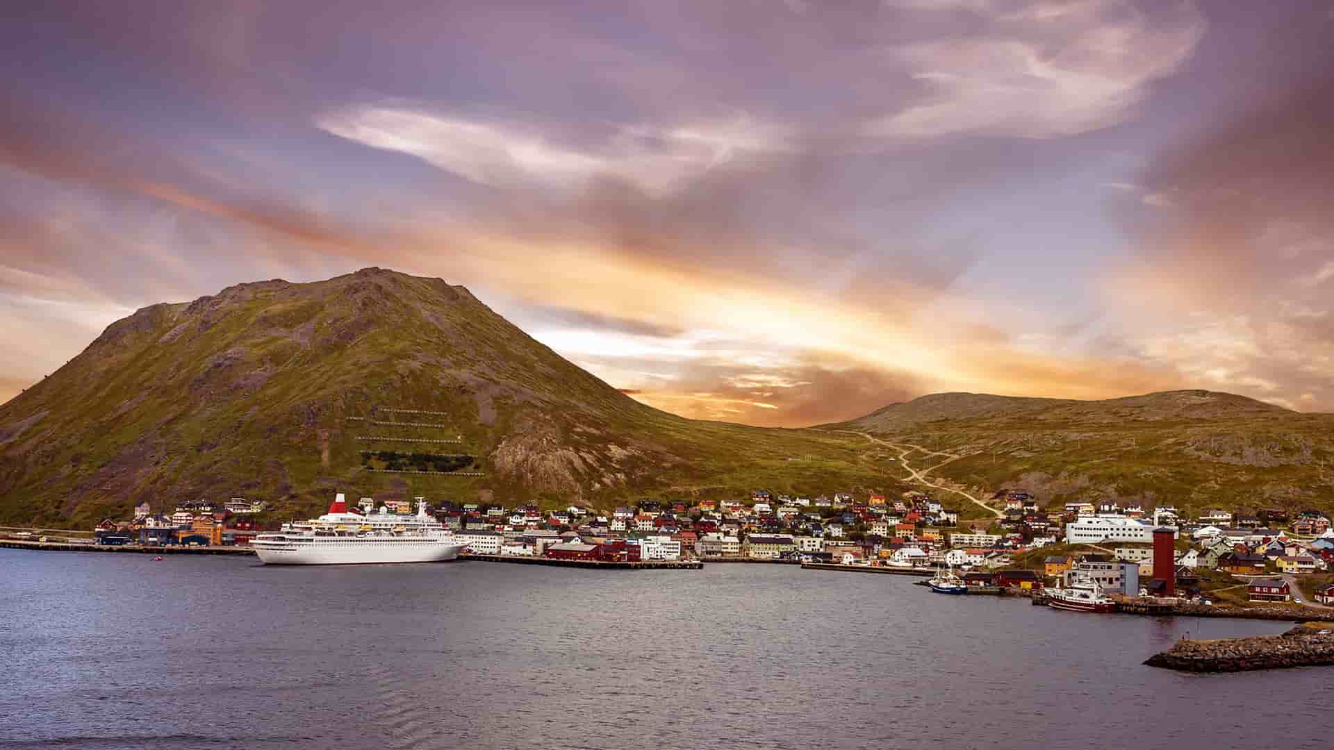 A large cruise ship is docked in the colorful harbor of Honningsvag, Norway, with the small town surrounded by a vast mountain landscape under a dramatic sky.