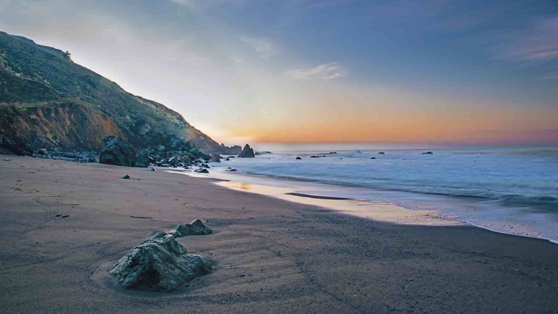A stunning sunset over a sandy beach with rocks and gentle waves, with a steep, grassy cliff on the left, at North Muiron Island, Australia.