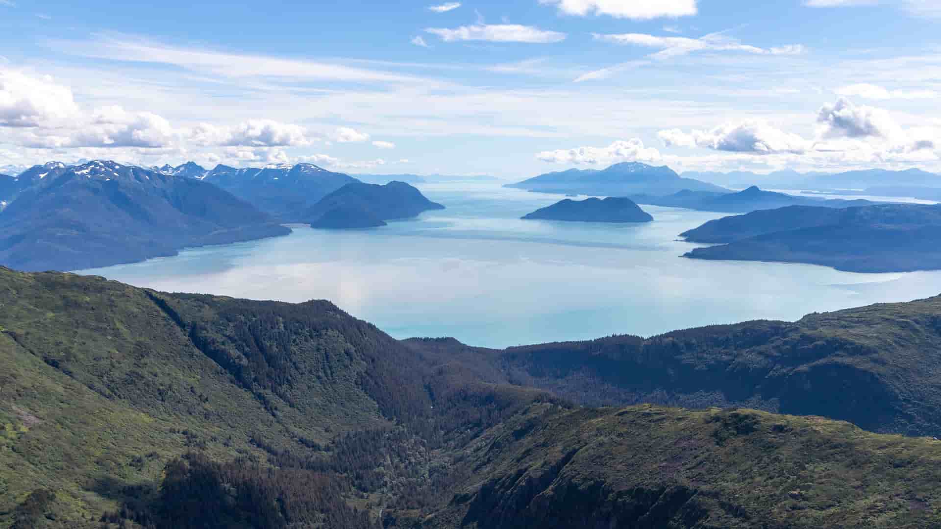 A scenic aerial view of the serene Northwest Passage, with turquoise water and numerous islands surrounded by majestic, snow-capped mountains under a blue sky with clouds.