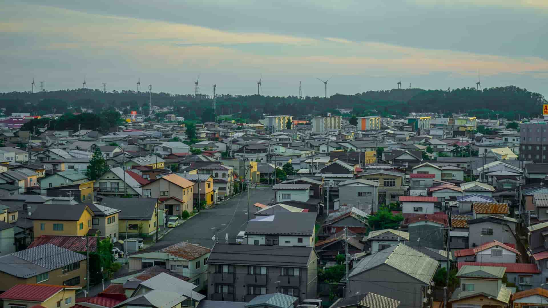 An elevated view over the dense residential neighborhood of Noshiro, Japan, with a line of wind turbines in the distance.