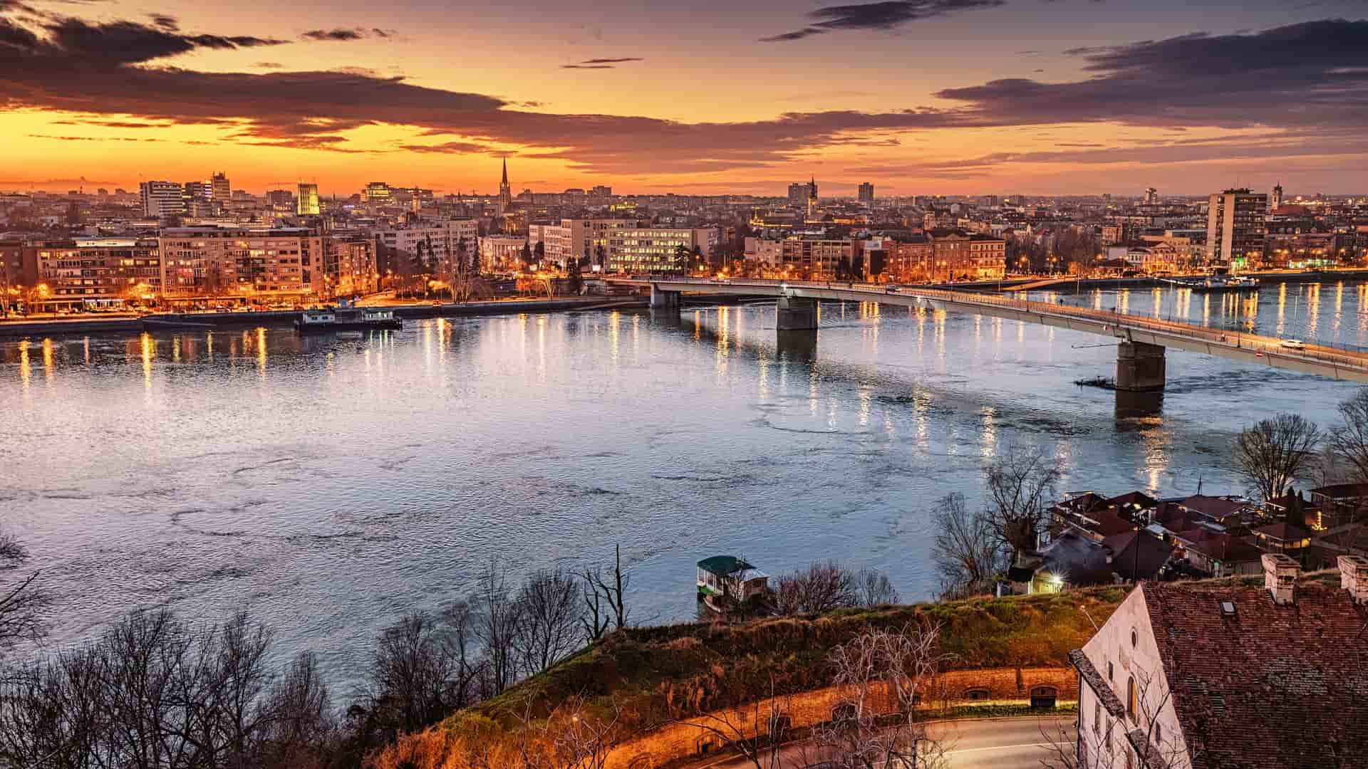 A wide-angle, elevated shot of the city of Novi Sad, Serbia, at dusk, showing the Danube River with bridges and city lights illuminating the skyline.