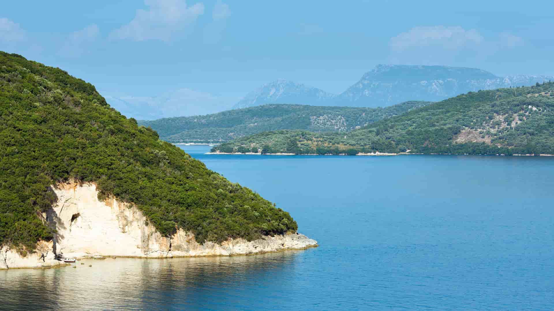 A beautiful daytime shot of the calm, blue waters and lush green hills of the bay of Nydri, Lefkada, with the Greek islands in the distance.