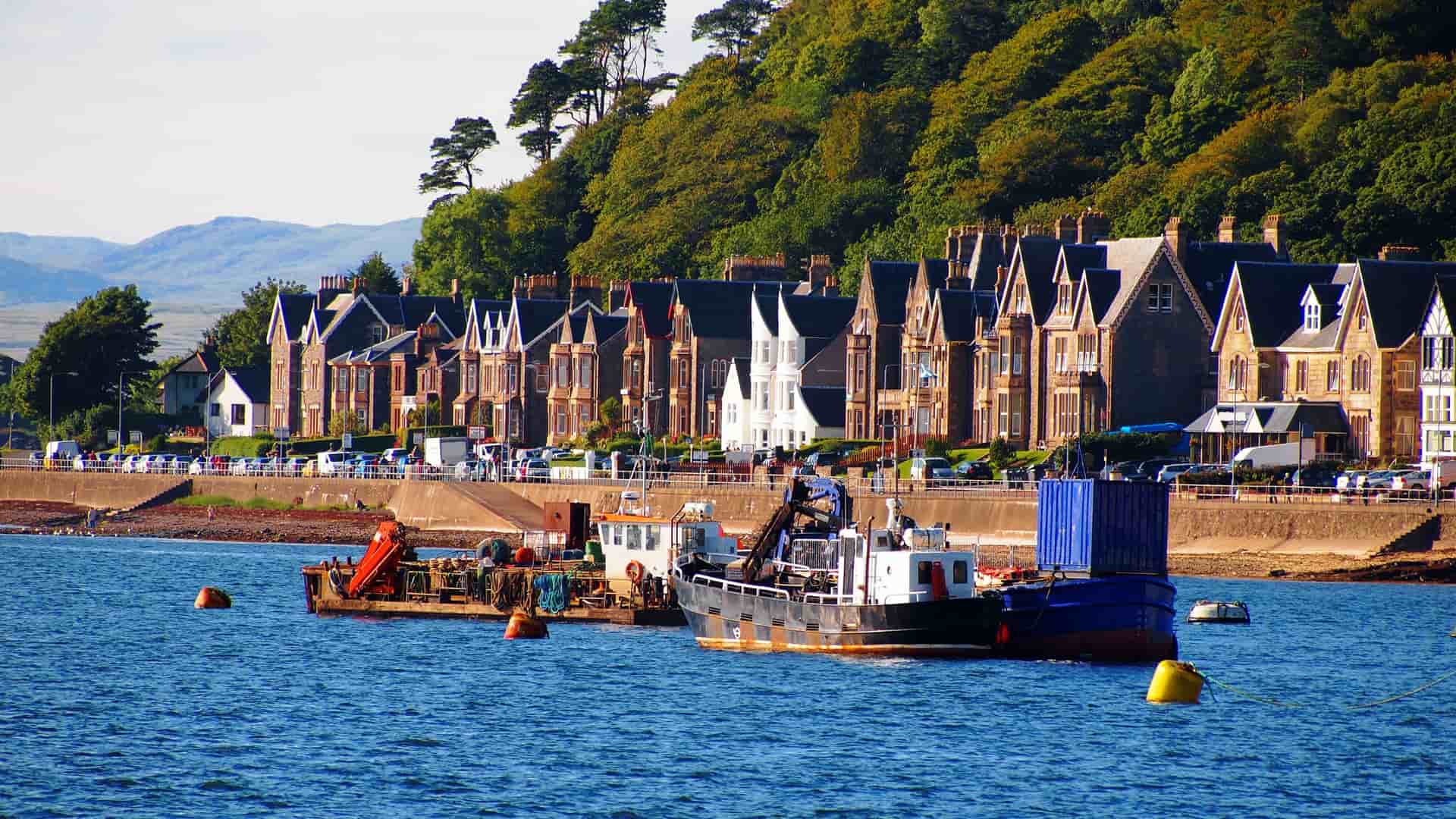 A vibrant shot of the waterfront in Oban, Scotland, with fishing boats docked in the bay in front of a row of Victorian-era townhouses and a lush green hillside.
