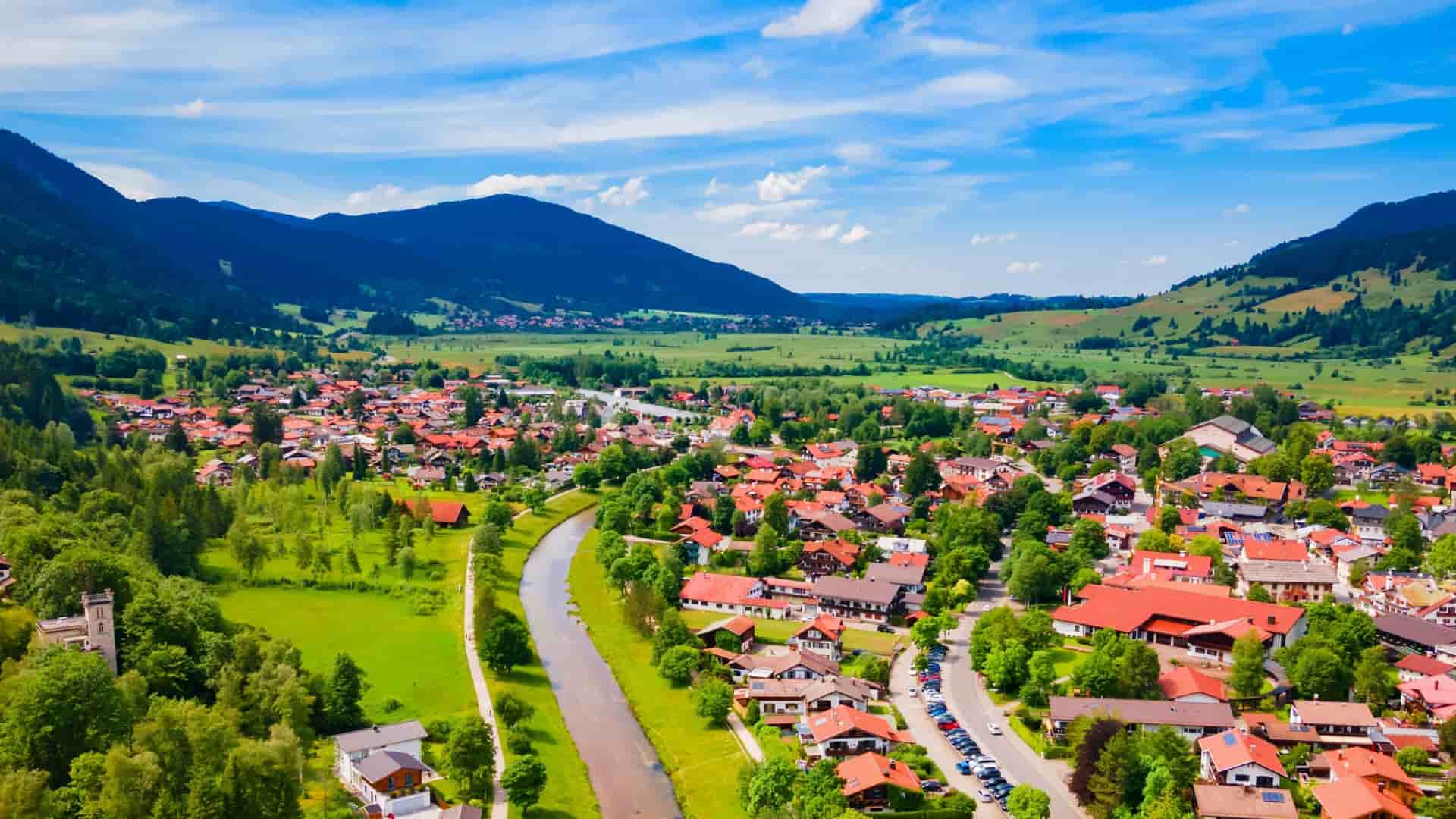 An aerial view of Oberammergau, a German village with red-roofed houses, situated in a green valley between rolling, forested mountains.