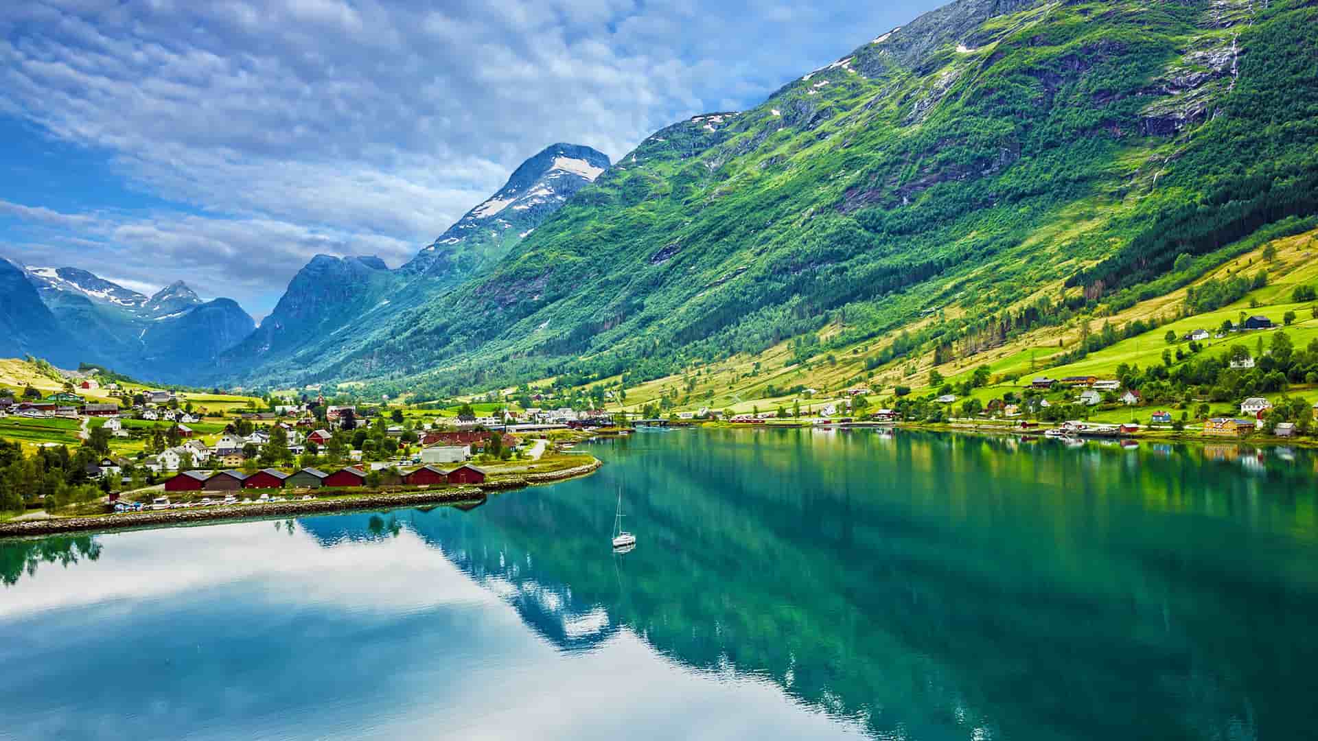 A scenic panoramic shot of the beautiful Olden village in Norway, with a peaceful lake reflecting the surrounding green mountains and scattered houses under a dramatic sky.