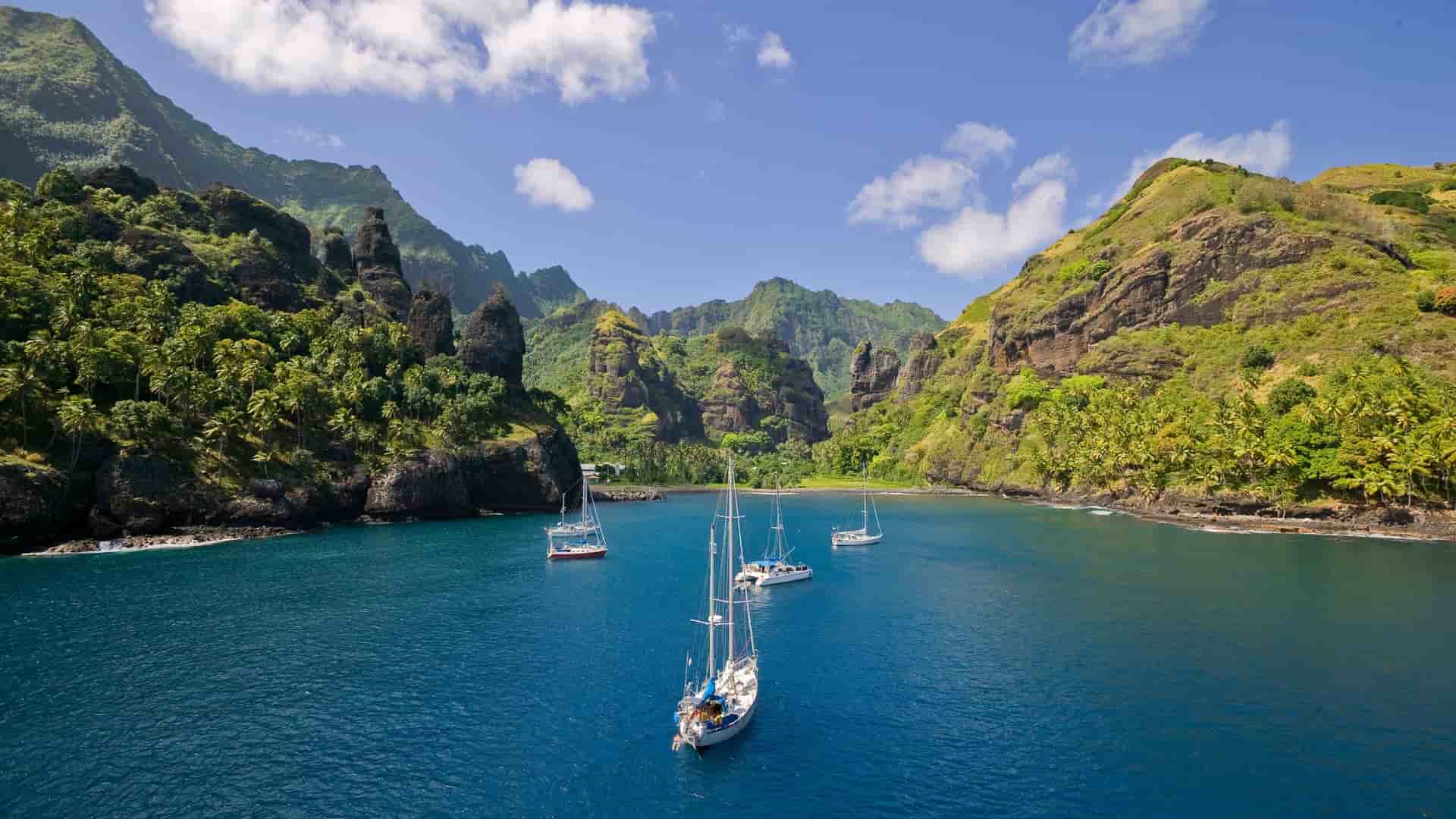 A beautiful scenic view of Omoa Bay on Fatu Hiva, Marquesas Islands, with several sailboats anchored in the peaceful blue water, surrounded by dramatic green cliffs and lush tropical vegetation.