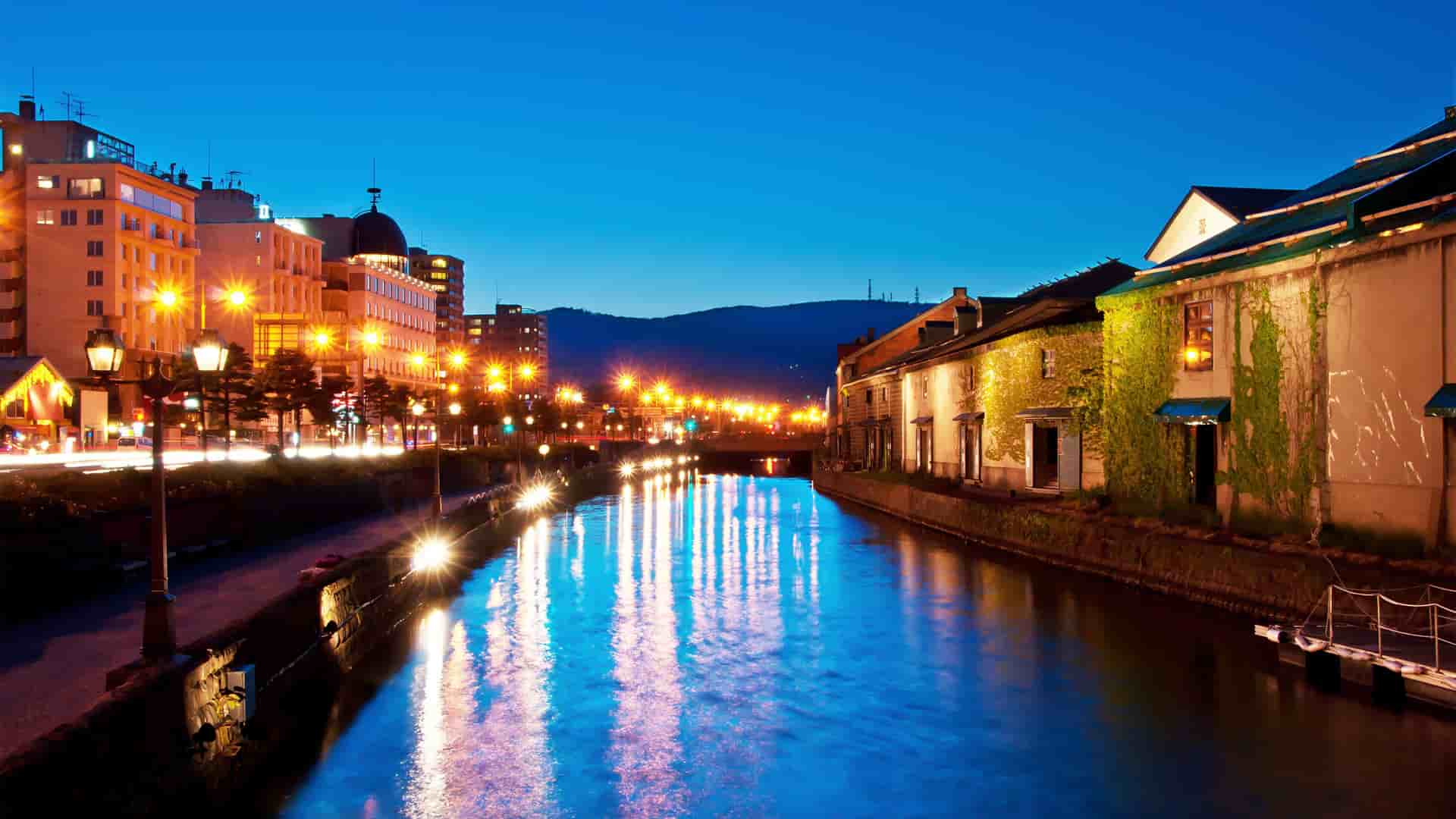 A stunning nighttime shot of the Otaru Canal in Japan, with historic warehouses and modern buildings illuminated on both sides, their lights reflecting beautifully on the water.