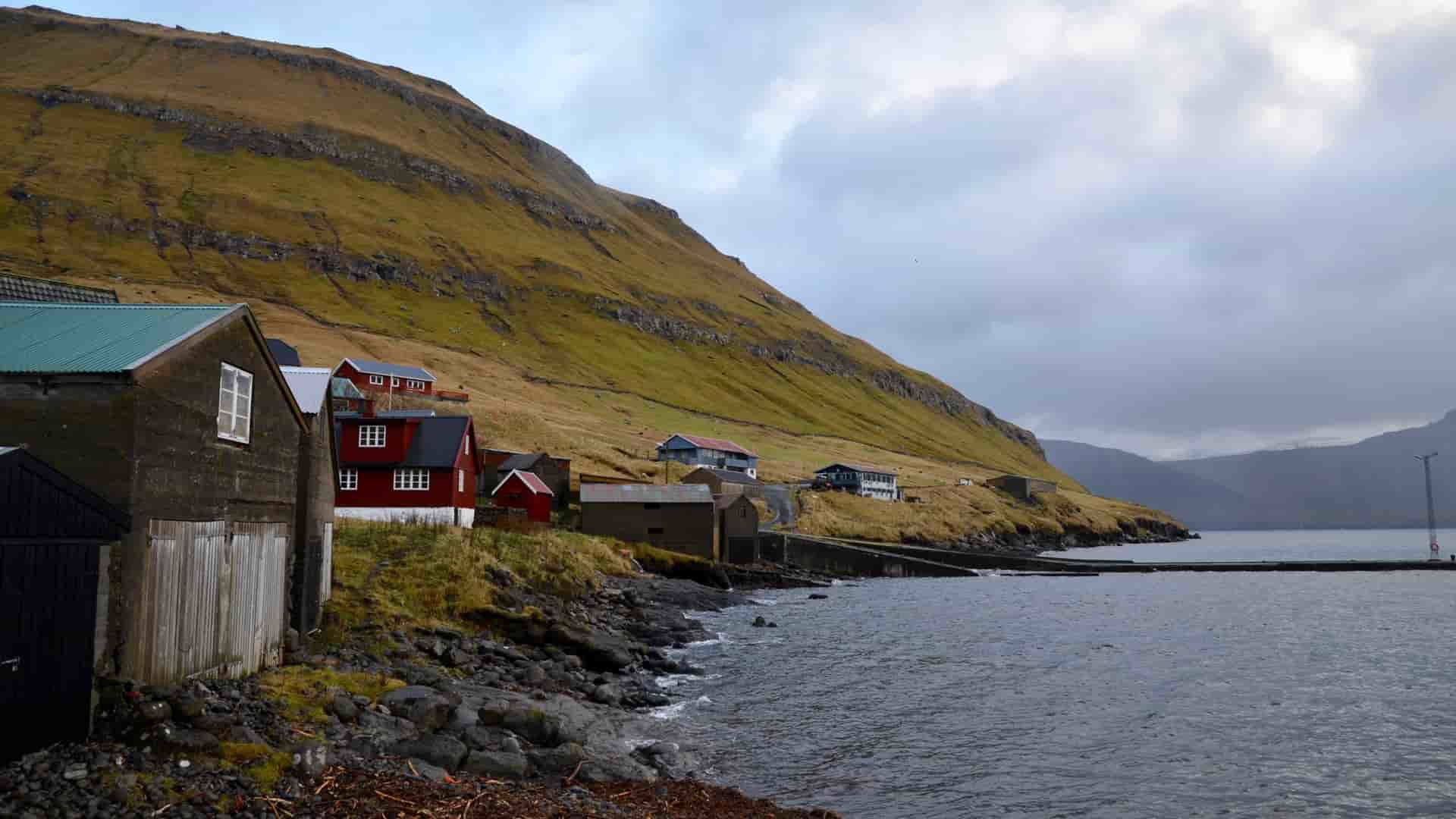 A view of a few houses with red and green roofs in the small, picturesque village of Oyndarfjørður in the Faroe Islands, nestled against a large green hill overlooking the harbor.