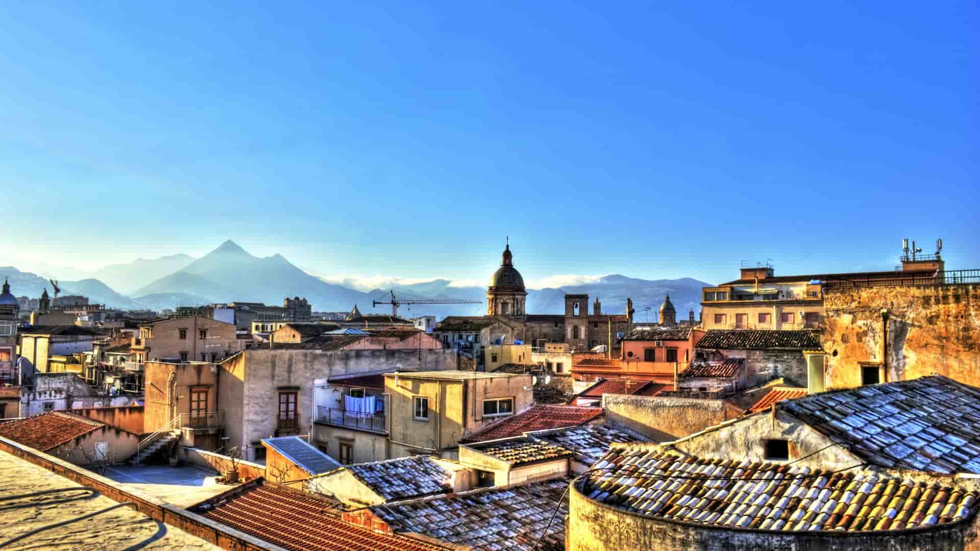 A high-angle view of the historic city of Palermo, Sicily, with a clear blue sky, buildings and rooftops in the foreground, and mountains in the distance.