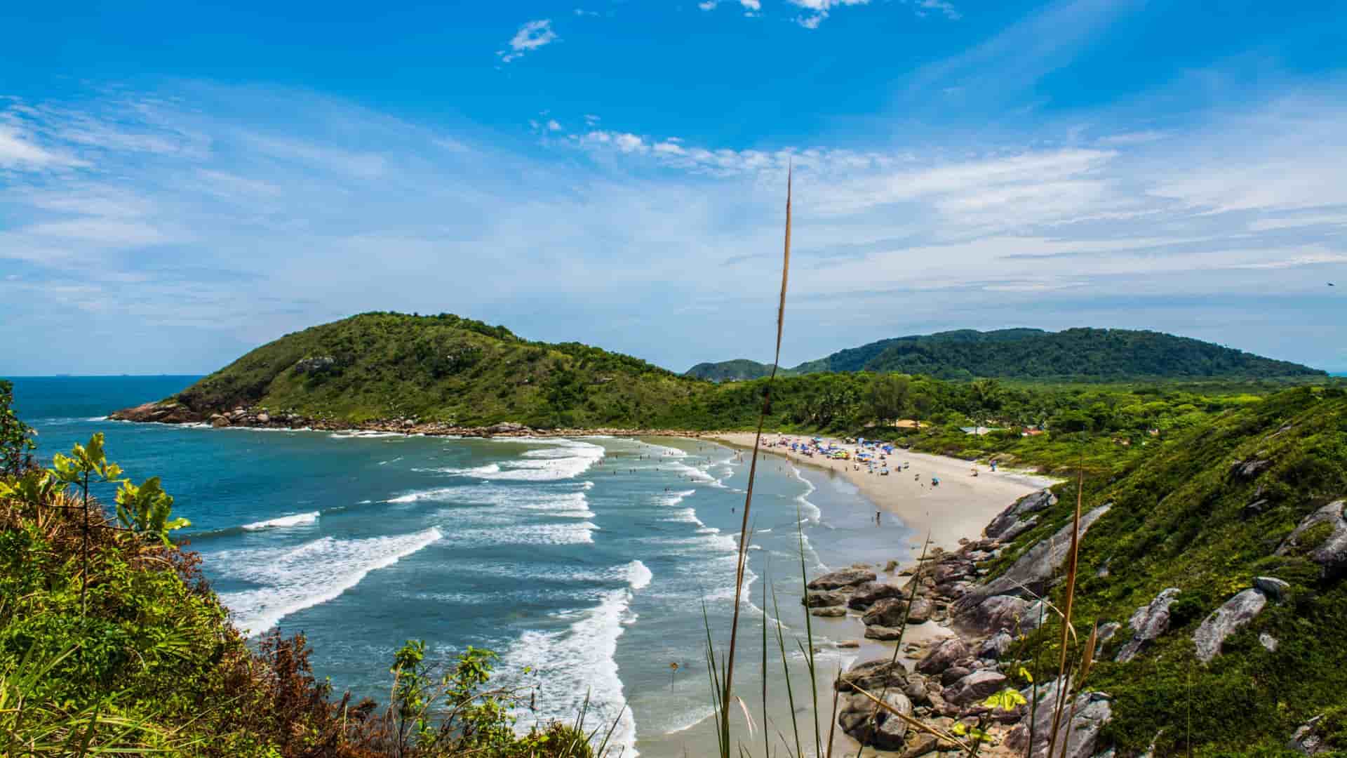 A panoramic aerial view of a beautiful sandy beach in the Bay of Paranaguá, Brazil, with waves crashing on the shore and lush, green hills in the distance.