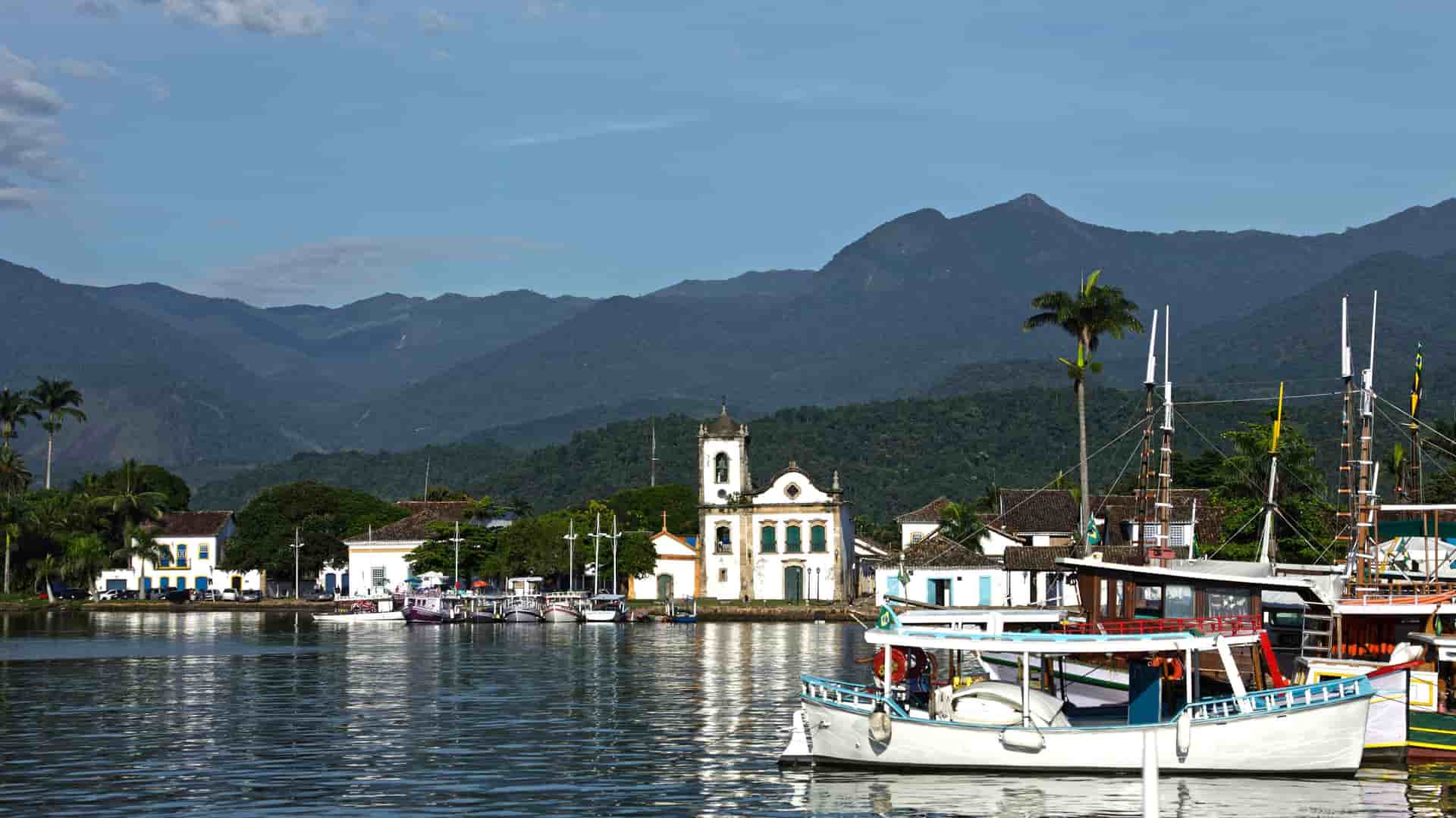 A view of the historic town of Paraty, Brazil, with boats docked in the harbor, colonial buildings, and mountains in the distance.