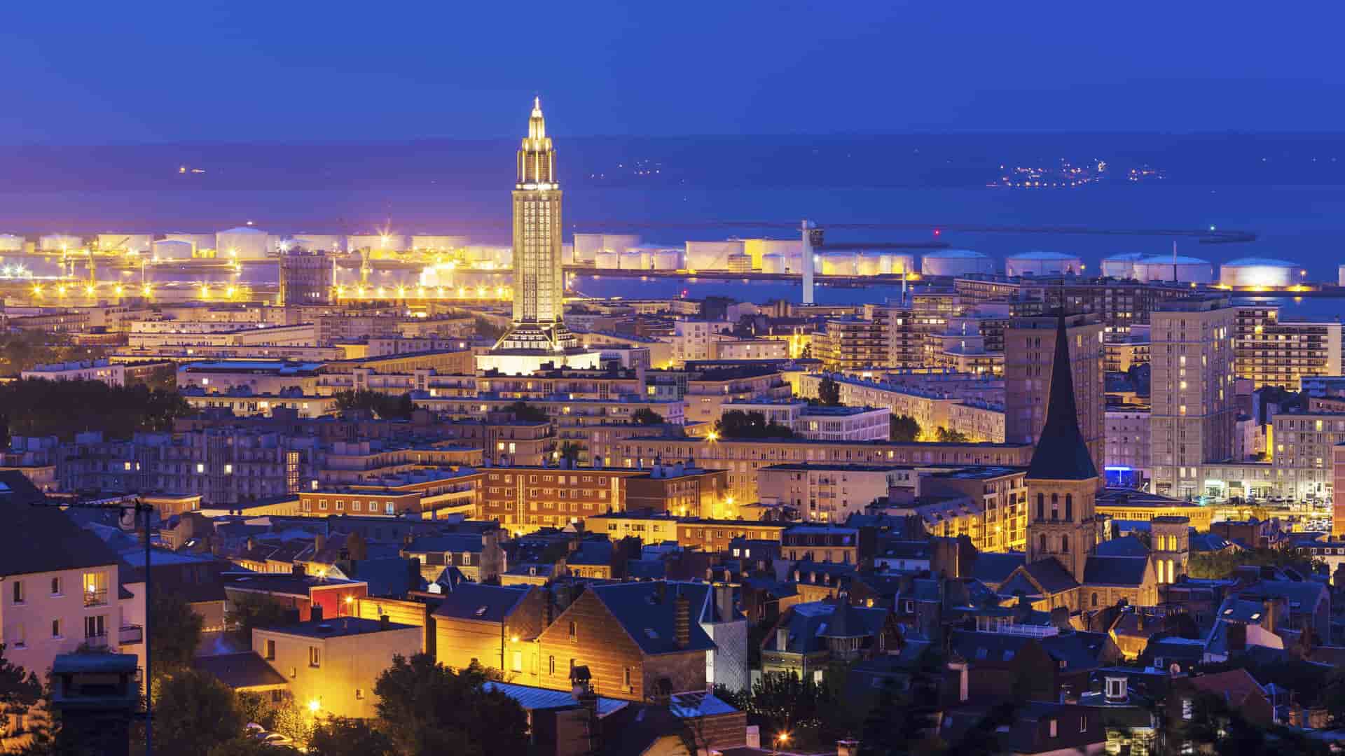 A wide-angle, nighttime view of Le Havre, France, with the tall Saint Joseph's Church steeple and the city lights illuminating the buildings and the harbor.