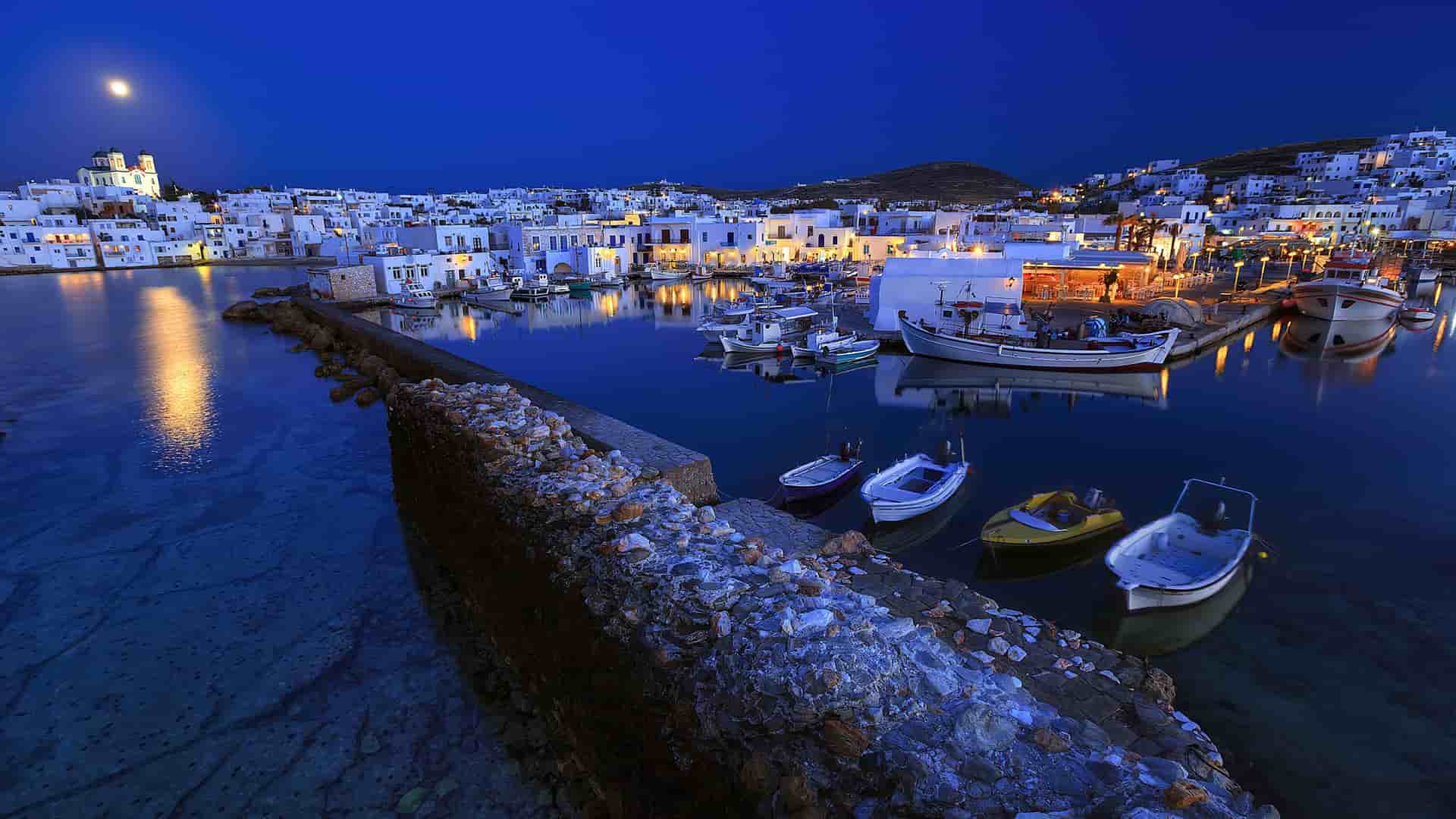 A nighttime shot of the Naoussa fishing harbor in Paros, Greece, with white buildings and boats docked in the calm water reflecting the lights and the moon.