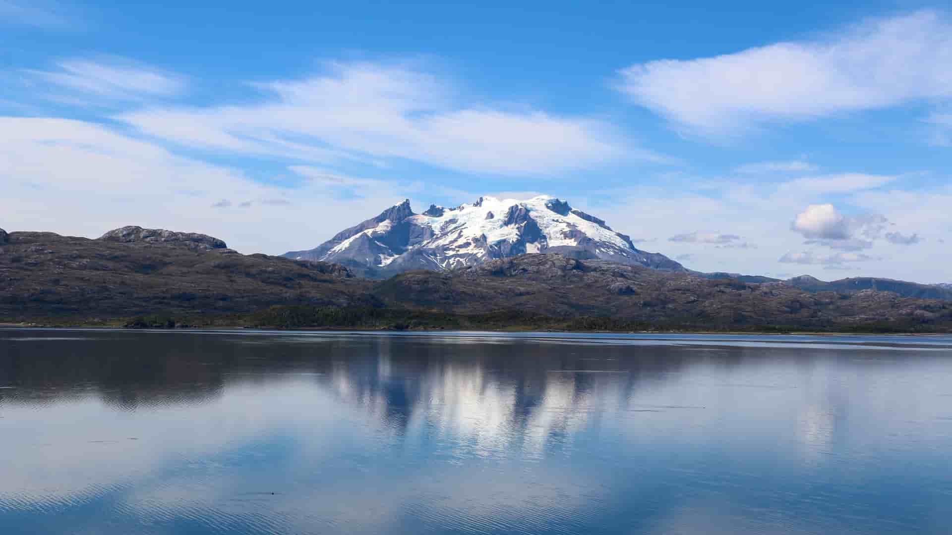 A scenic view of Peel Fjord in Chile, with a snow-capped mountain reflecting on the calm water and a cloudy blue sky in the background.