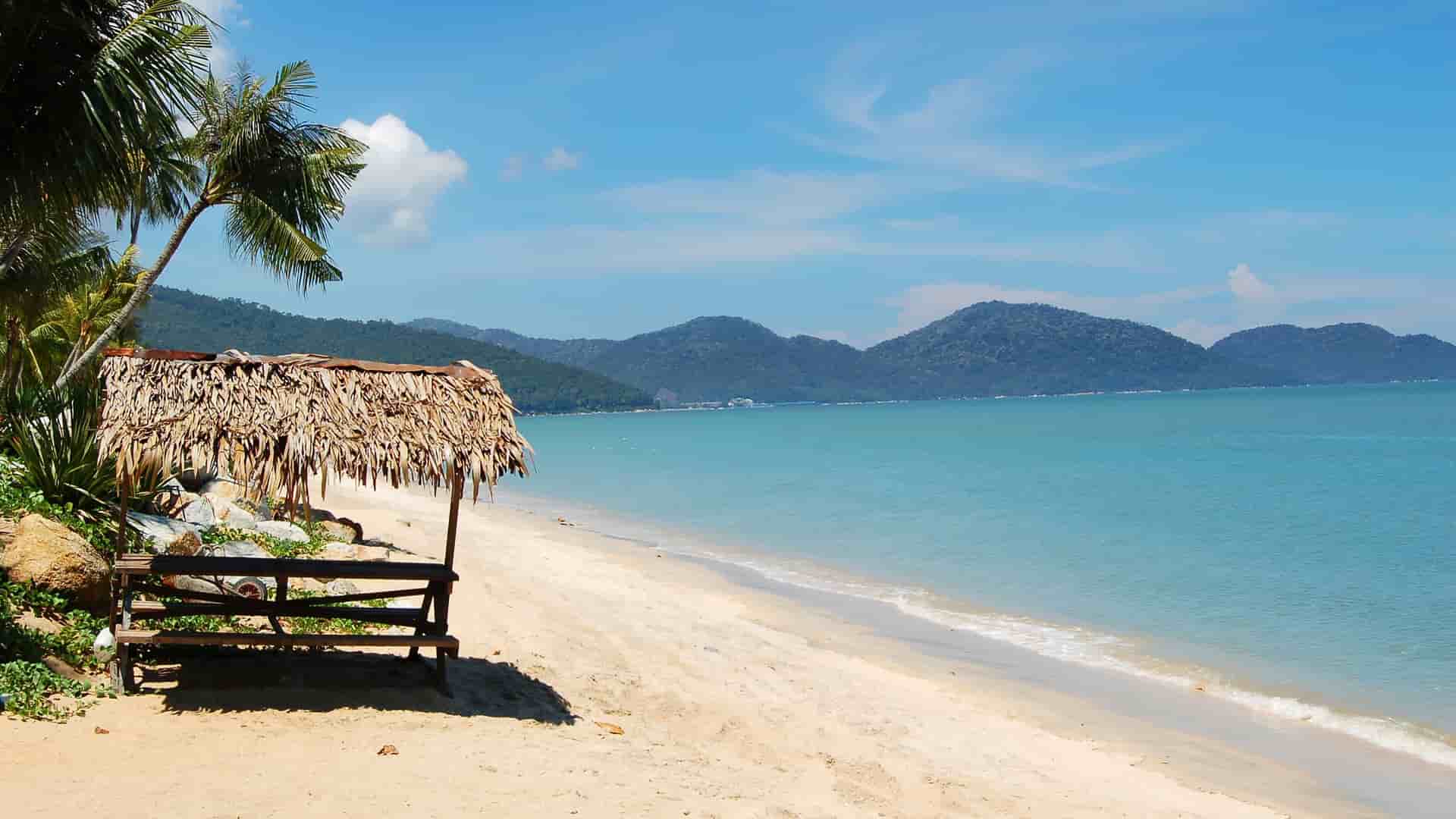 A view of the sandy beaches of Penang, Malaysia, with a thatched-roof hut on the shore, palm trees, and the clear blue ocean.