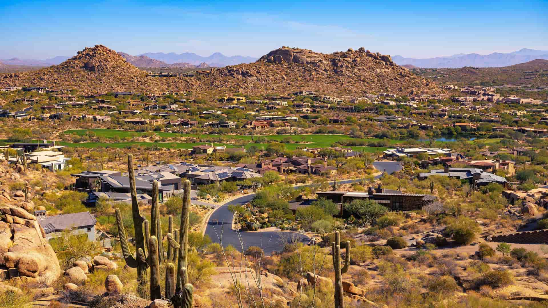 A panoramic view of a residential neighborhood in the desert hills near Phoenix, Arizona, with a golf course visible among the homes. The foreground features saguaro cacti and desert plants, while a winding road cuts through the landscape, leading towards the rocky mountains under a clear blue sky.