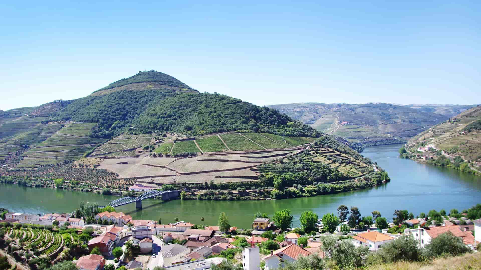 A panoramic view of the Douro River in Portugal, showcasing the terraced vineyards on the hillsides surrounding the small town of Pinhão, with a bridge crossing the water.