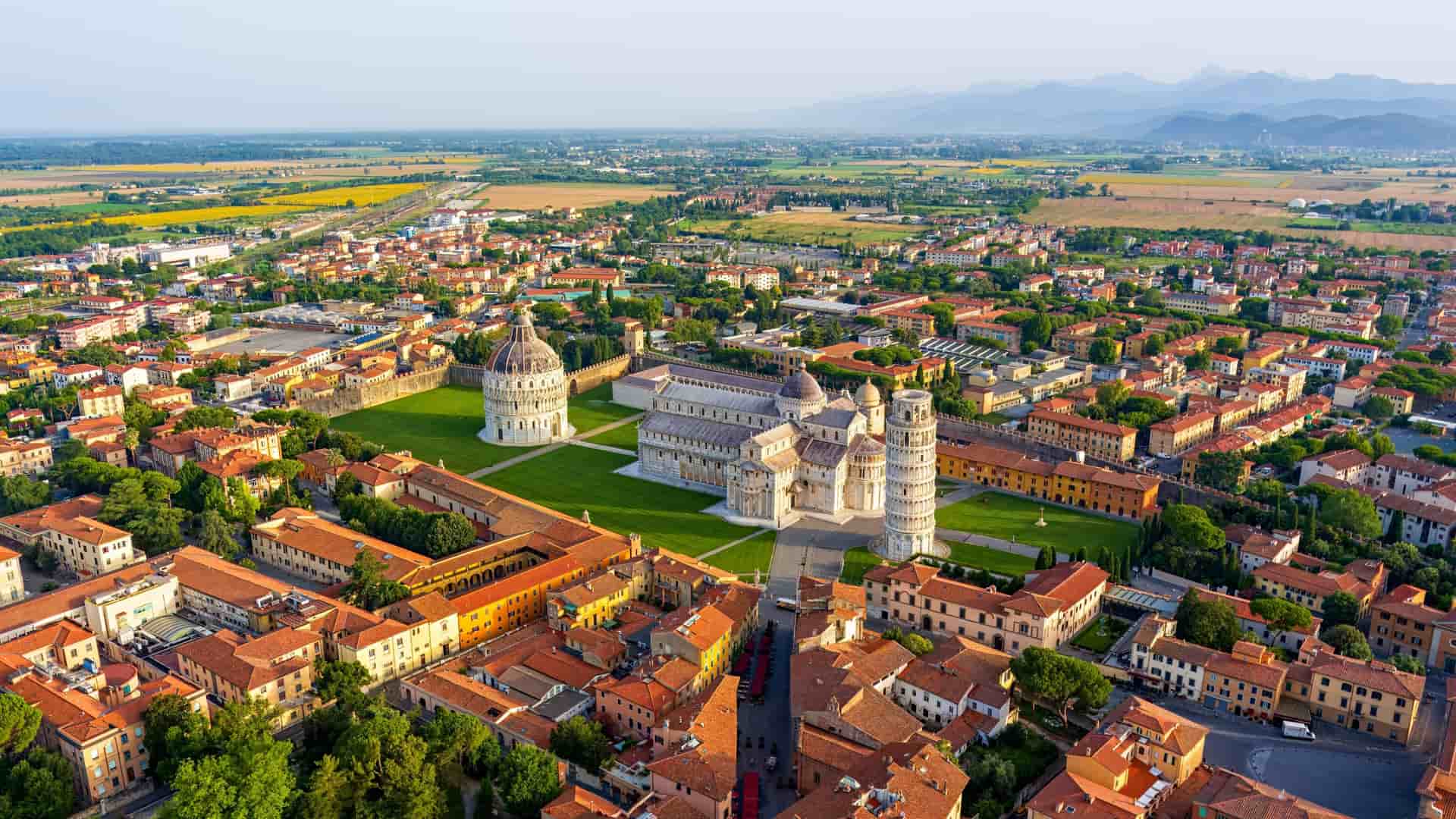 High aerial view of the Leaning Tower, Cathedral, and Baptistery on a lush green lawn, surrounded by the red-roofed buildings of Pisa.