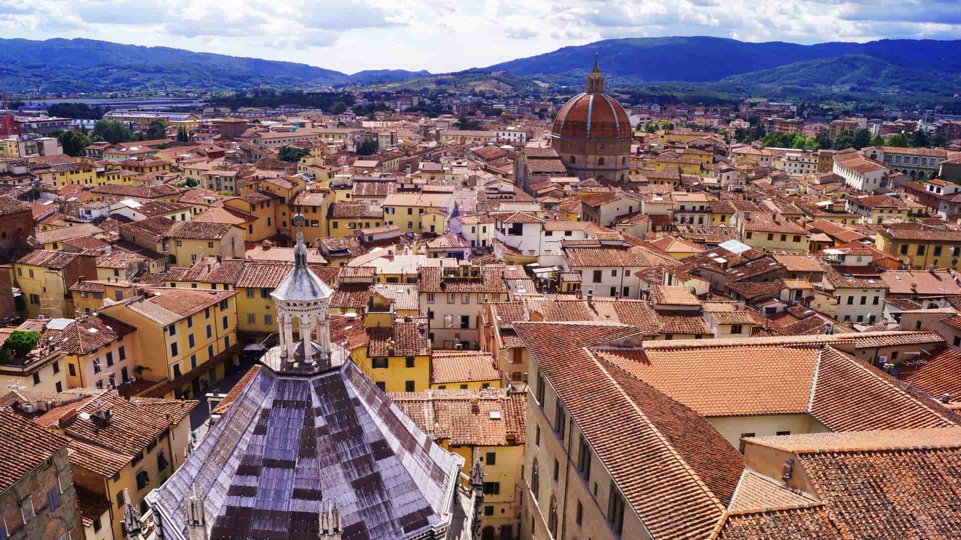 An aerial view of the historic city center of Pistoia, Tuscany, Italy, showcasing the tightly packed terracotta rooftops of medieval and Renaissance buildings. The prominent feature is the large, octagonal dome of the Basilica of Santa Maria dell'Umiltà, rising above the surrounding cityscape.