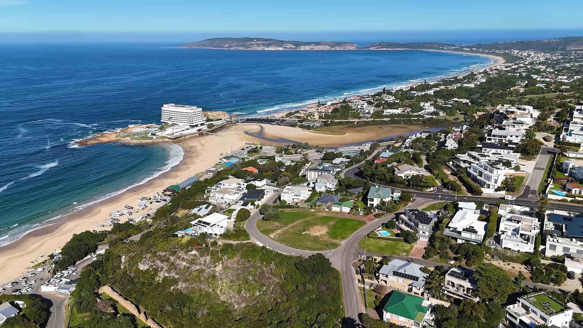 An aerial view of Plettenberg Bay, South Africa, showing a sweeping coastline with a wide sandy beach. The town's white houses and buildings are nestled on the green hills that slope down to the bay, where a river empties into the ocean. A prominent headland is visible in the distance under a clear blue sky.