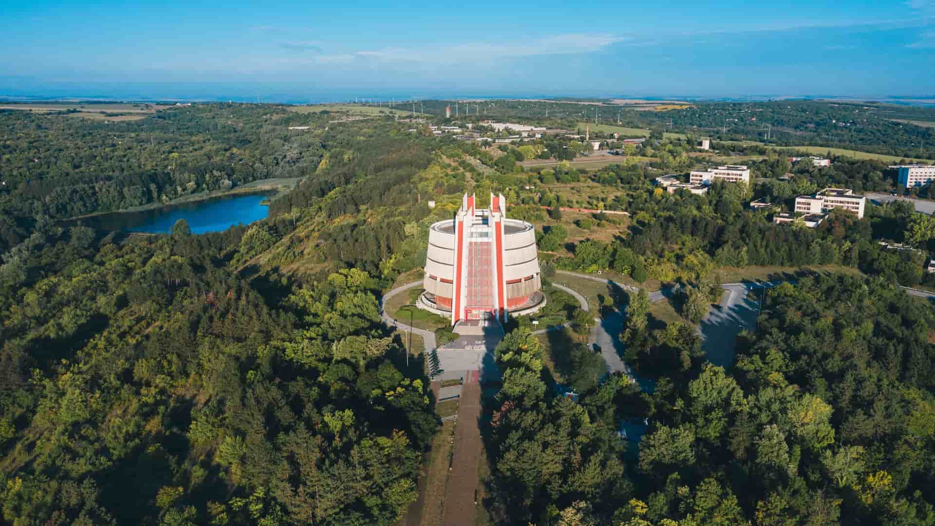An aerial view of the iconic Pleven Panorama in Bulgaria, a modern, cylindrical building surrounded by dense green forests and a lake, commemorating the Siege of Pleven.