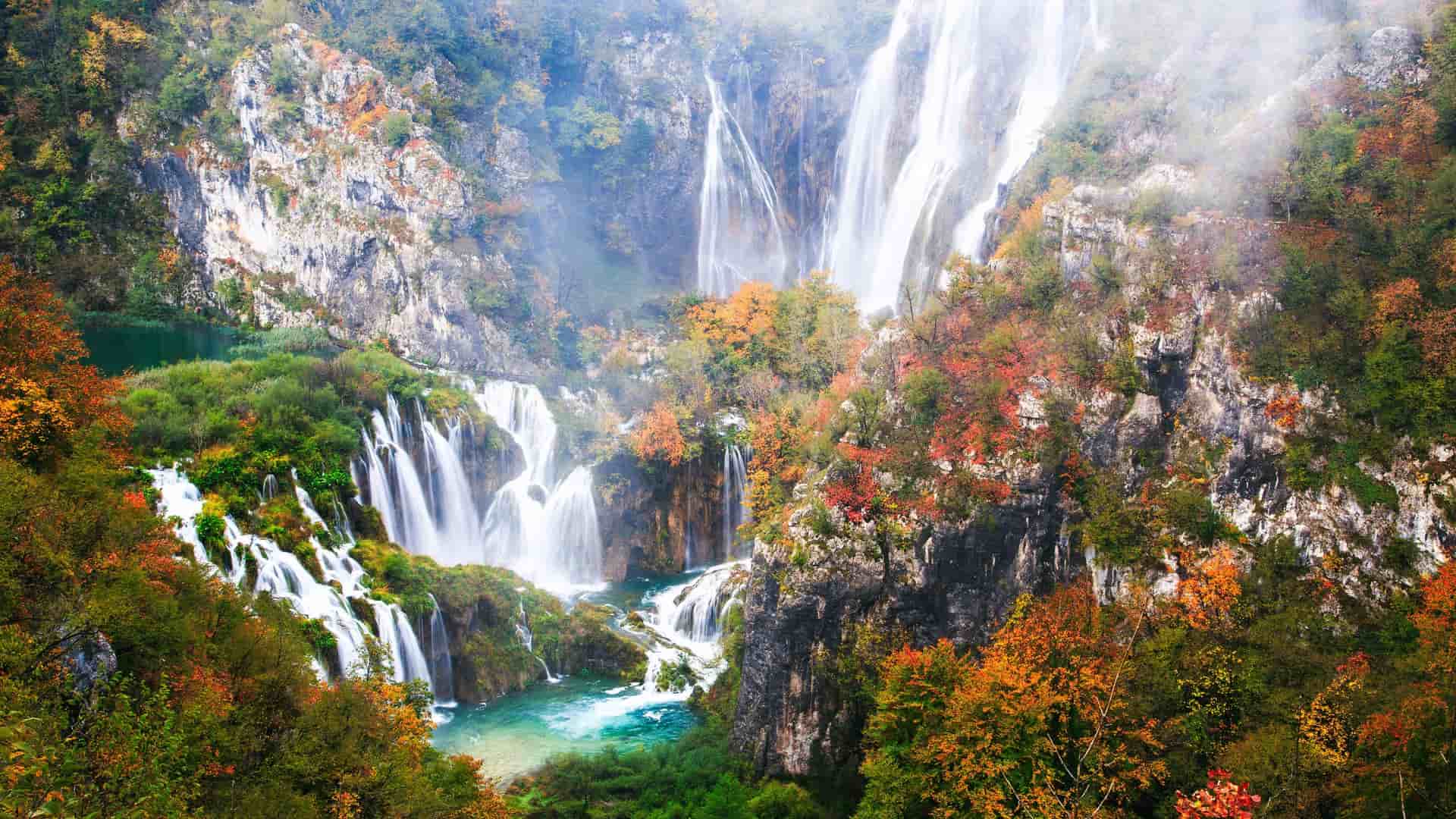A breathtaking view of the Plitvice Lakes National Park in Croatia, showing multiple waterfalls cascading down rugged cliffs and into vibrant turquoise pools. The surrounding forest is in beautiful autumn colors of orange, red, and green, with a light mist rising from the water.