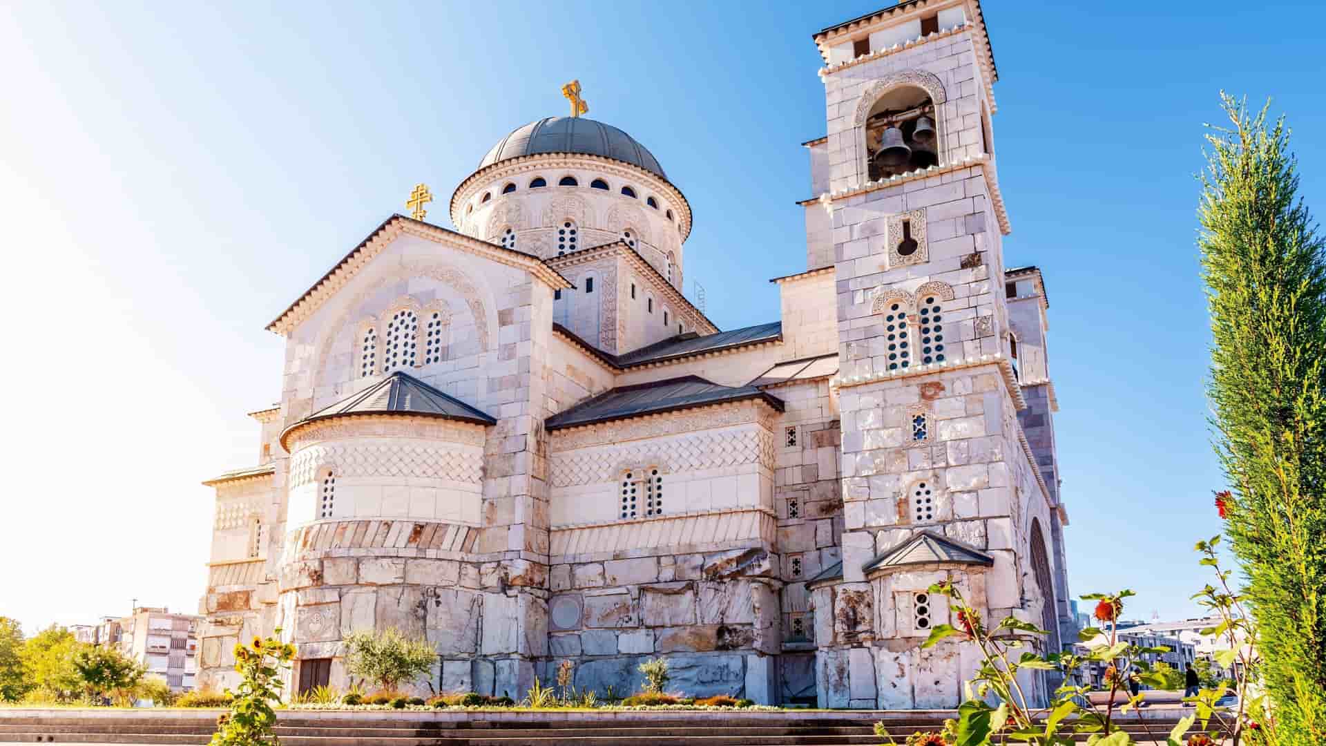 A close-up view of the Resurrection of Christ Orthodox Cathedral in Podgorica, Montenegro, highlighting its intricate white stone facade, domed roof, and a separate bell tower under a clear blue sky.