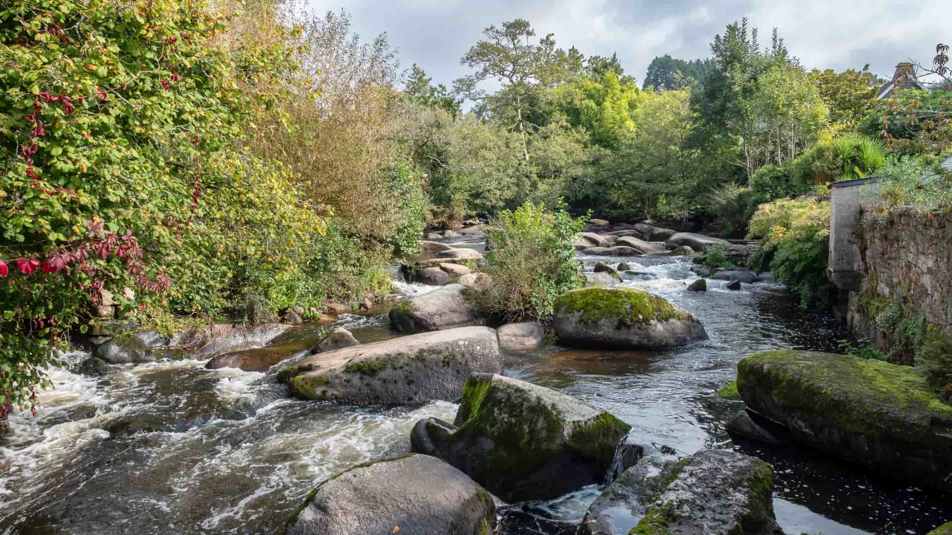 A picturesque view of the Aven River flowing through Pont-Aven, France, with large moss-covered boulders in the shallow, rushing water and trees on the banks.
