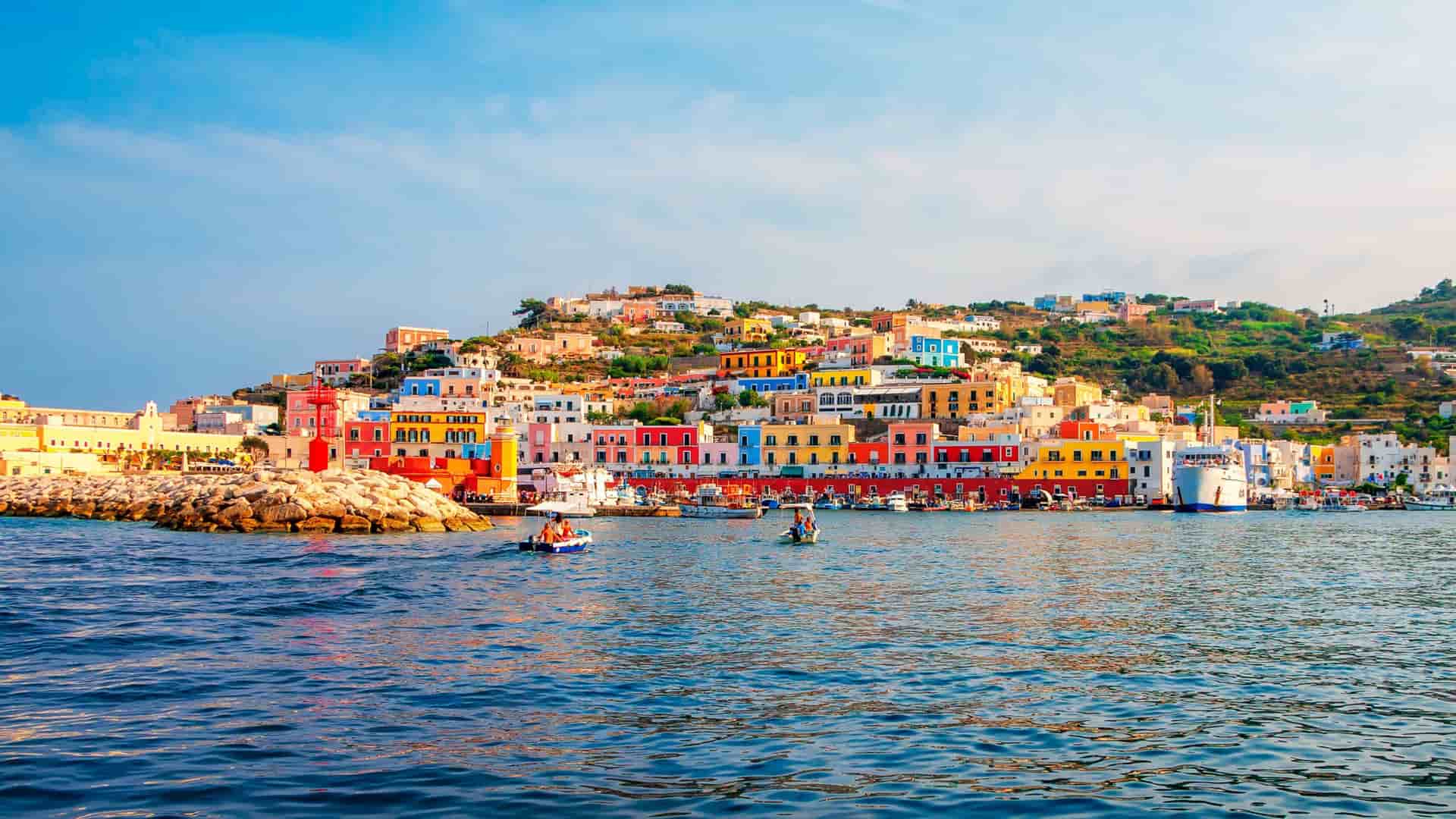 A colorful view of the port of Ponza Island in Italy, with brightly painted houses stacked on the hillside overlooking the blue Tyrrhenian Sea and boats docked along the waterfront.