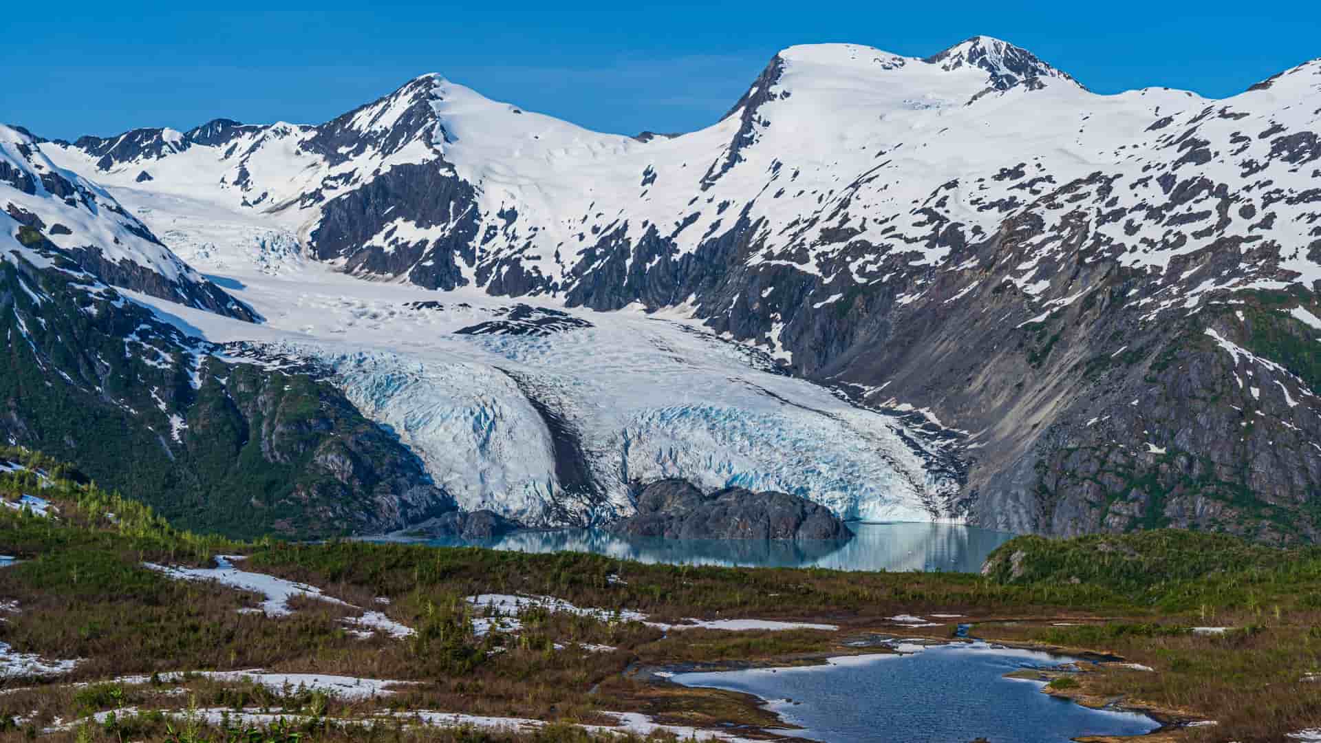 A vast, blue-ice glacier flows down between two snow-capped mountains and ends in a dark, clear lake below.