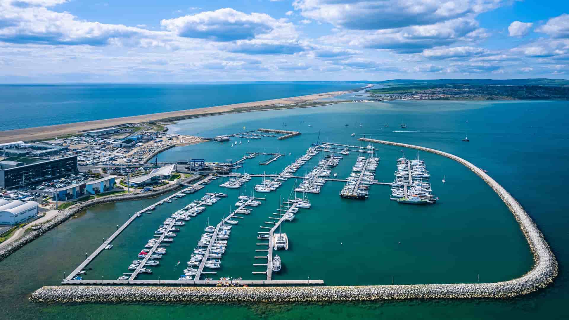 An aerial view of Portland Harbour in Dorset, England, showcasing a large marina filled with docked sailboats and yachts, a long stone breakwater, and the sweeping Chesil Beach in the distance under a blue sky with scattered clouds.