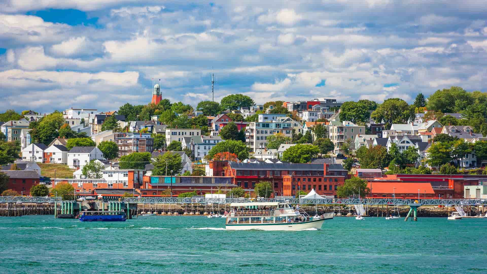 A panoramic view of the city of Portland, Maine, with a colorful waterfront and a variety of boats sailing on the water in the foreground and the city buildings and a lighthouse on the hill in the background.