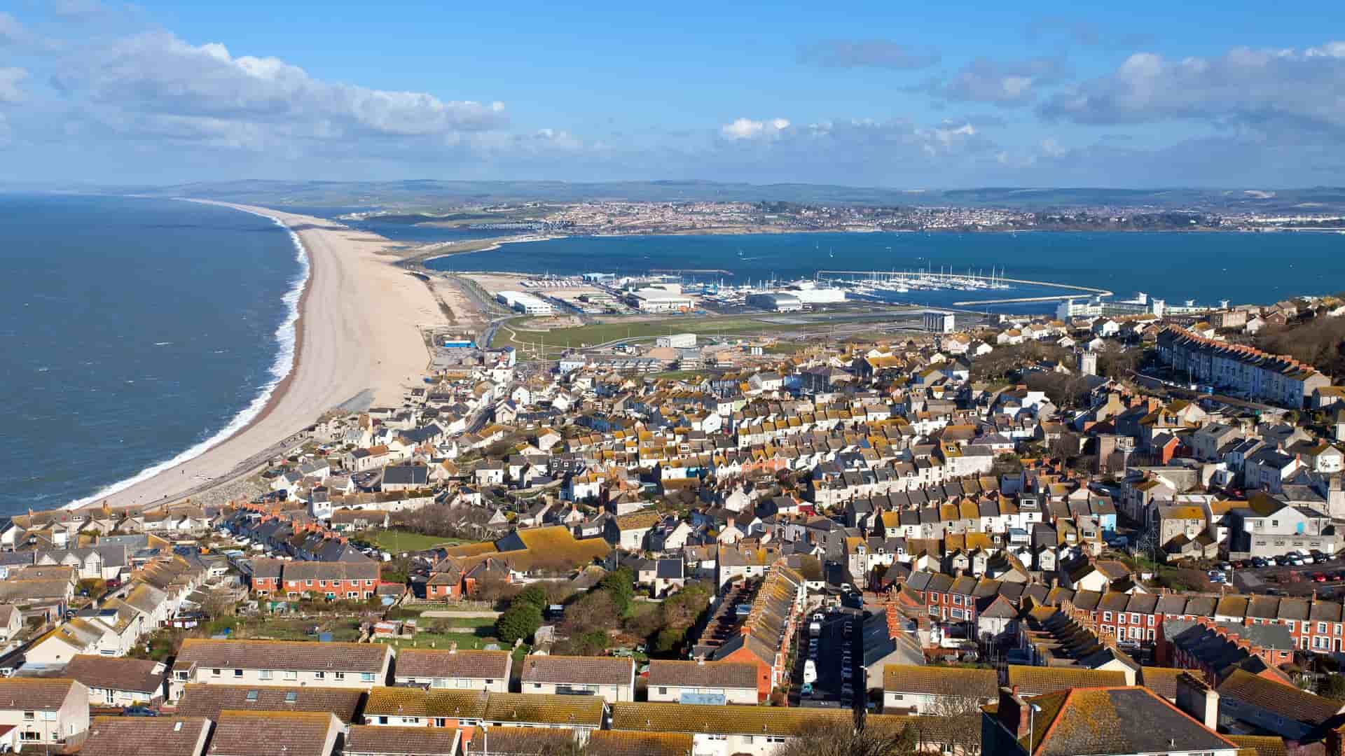 An aerial view of the seaside town of Portland in Dorset, England, showcasing the densely packed rooftops, the vast expanse of Chesil Beach, and the Portland Harbour in the background.
