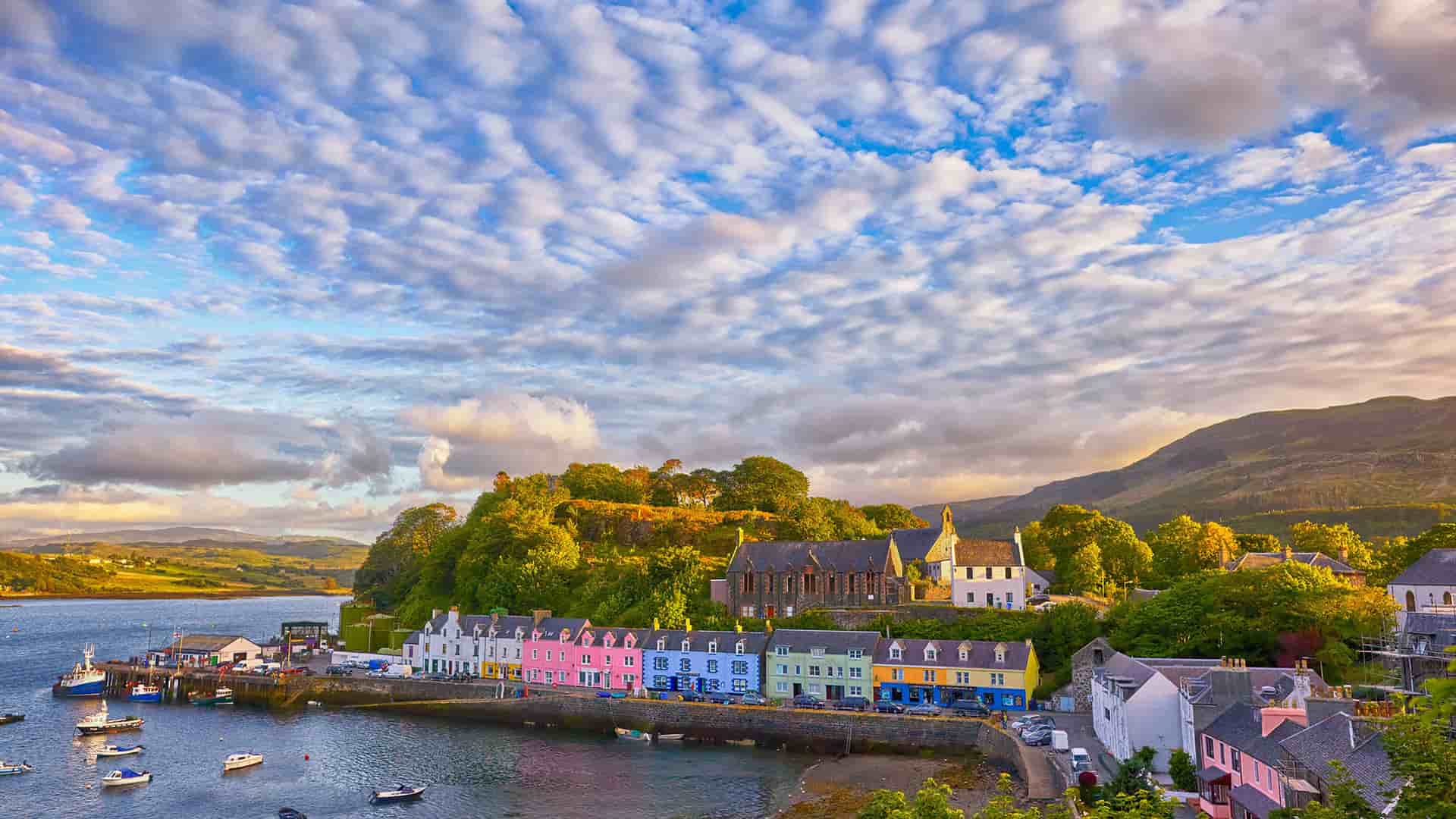 A stunning sunrise over the picturesque harbor of Portree on the Isle of Skye, Scotland, with its iconic colorful waterfront buildings and fishing boats under a dramatic sky.