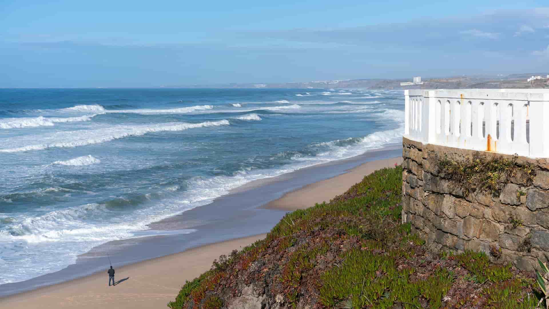 A serene view of Praia de Santa Cruz in Portugal, with large ocean waves rolling onto a wide, sandy beach. In the foreground, a stone wall with a white balustrade sits on a grassy cliff, while a lone person is fishing on the shore below. The sky is partly cloudy.