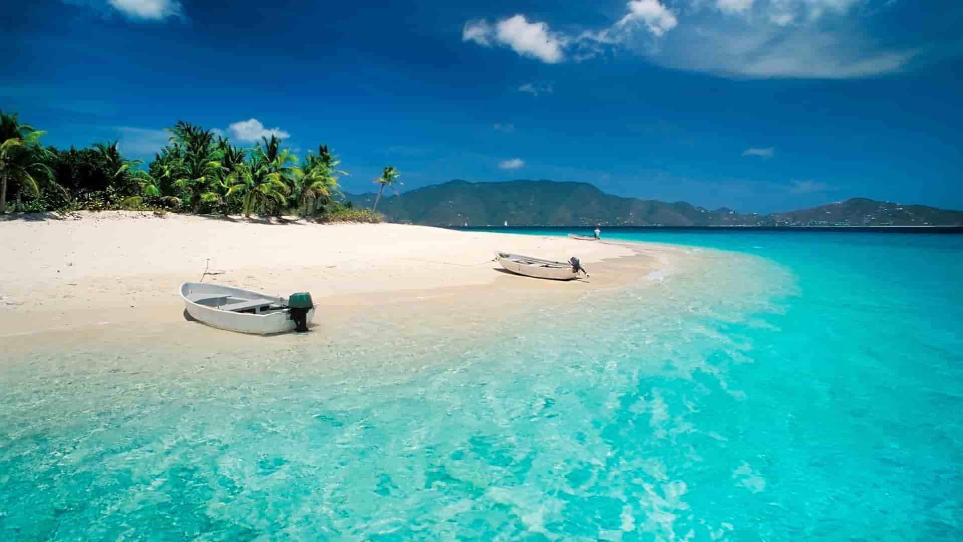 A bright tropical scene in Prickly Pear Island BVI with clear turquoise water, white sand, two small boats pulled onto the beach, and lush palm trees set against distant green mountains under a vivid blue sky.