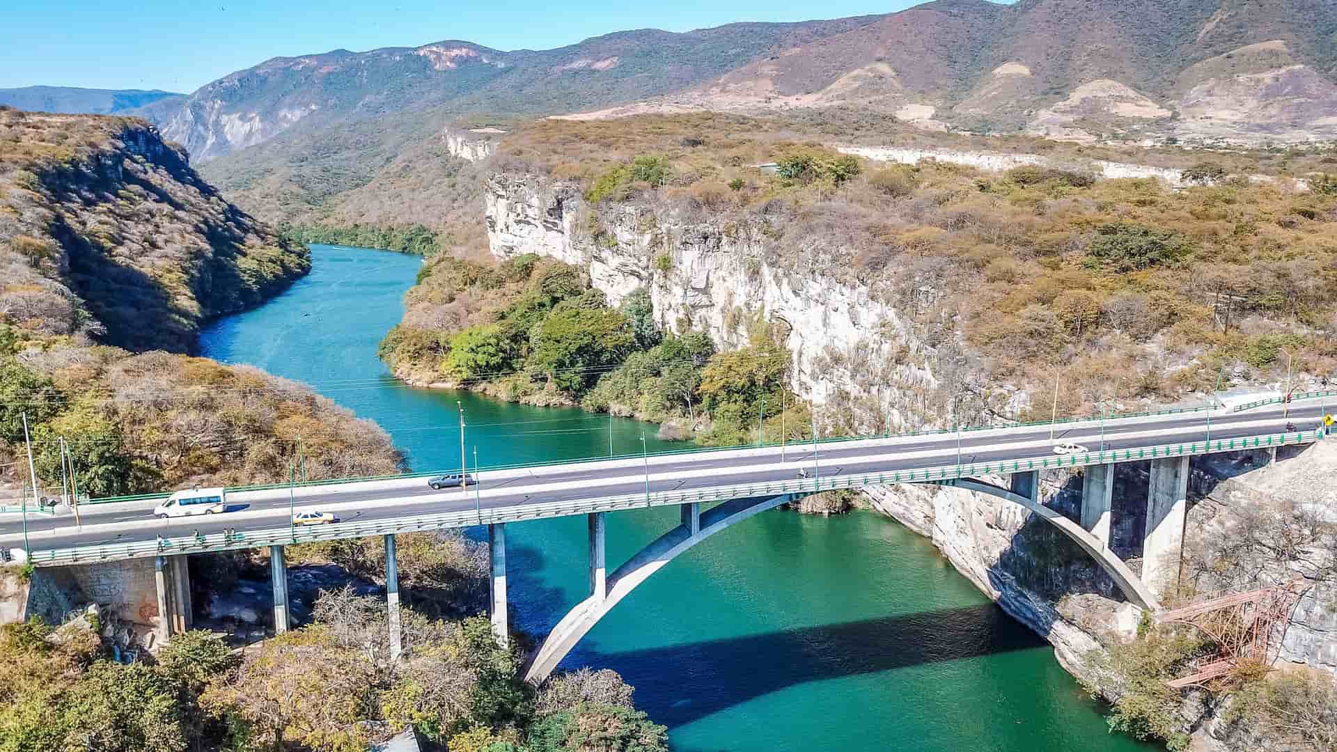 A wide-angle aerial view of the Puente Belisario Domínguez bridge spanning the turquoise Grijalva River in Sumidero Canyon, Chiapas, Mexico. The bridge is surrounded by steep, rocky cliffs and mountains with arid vegetation.