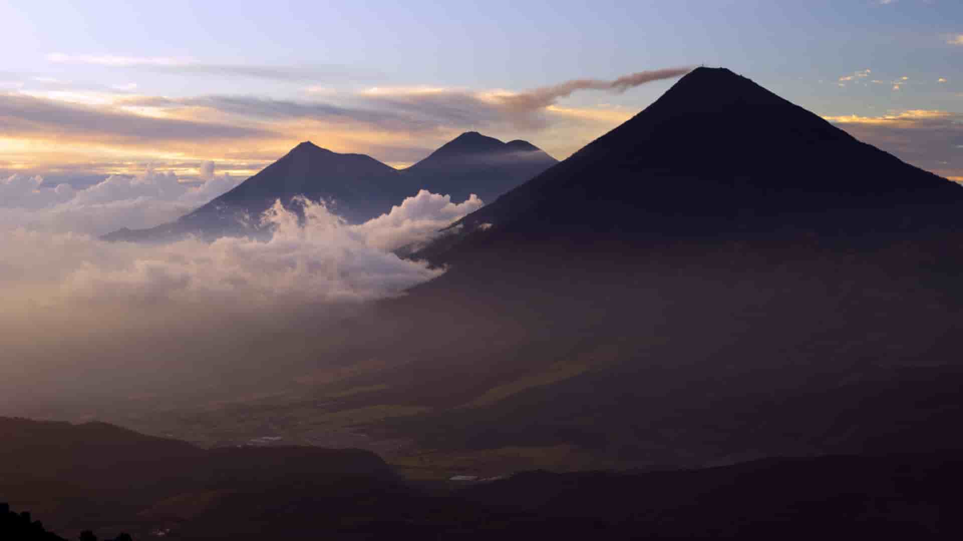 A wide-angle landscape shot of the three iconic volcanoes, Agua, Fuego, and Acatenango, in Guatemala, rising above the clouds during a vibrant sunrise, a scenic view near Puerto Quetzal.