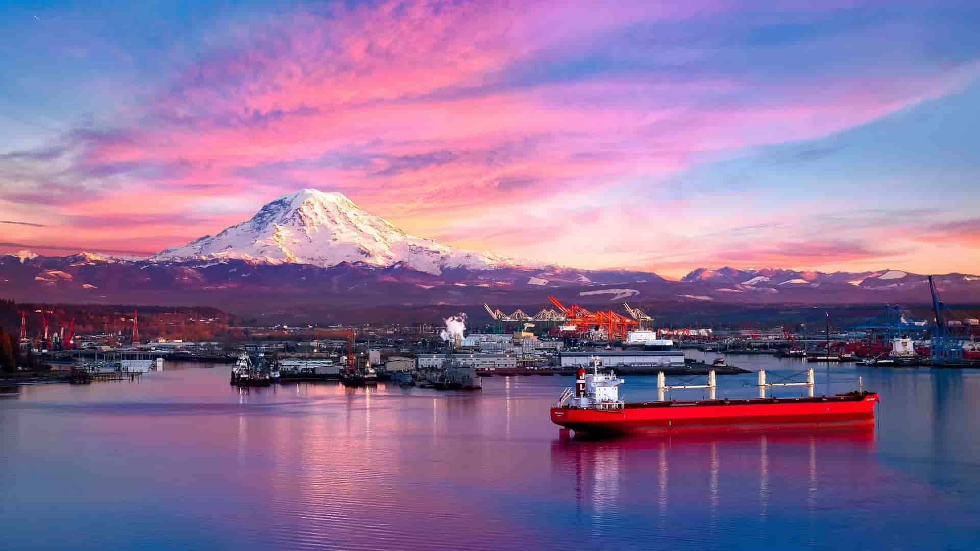 A vibrant sunset over Commencement Bay and the port of Tacoma in Puget Sound, Washington, with a red cargo ship in the foreground and the majestic snow-capped Mount Rainier in the distance.