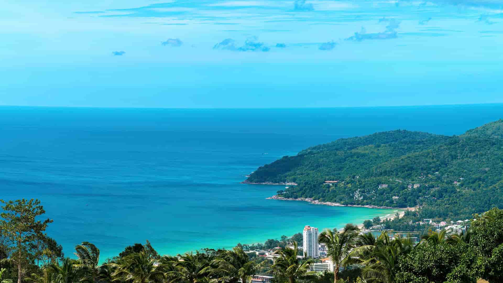 A stunning high-angle shot from a viewpoint overlooking Nai Harn Beach in Phuket, Thailand, showing a beautiful bay with turquoise water and lush green hills stretching into the deep blue ocean.
