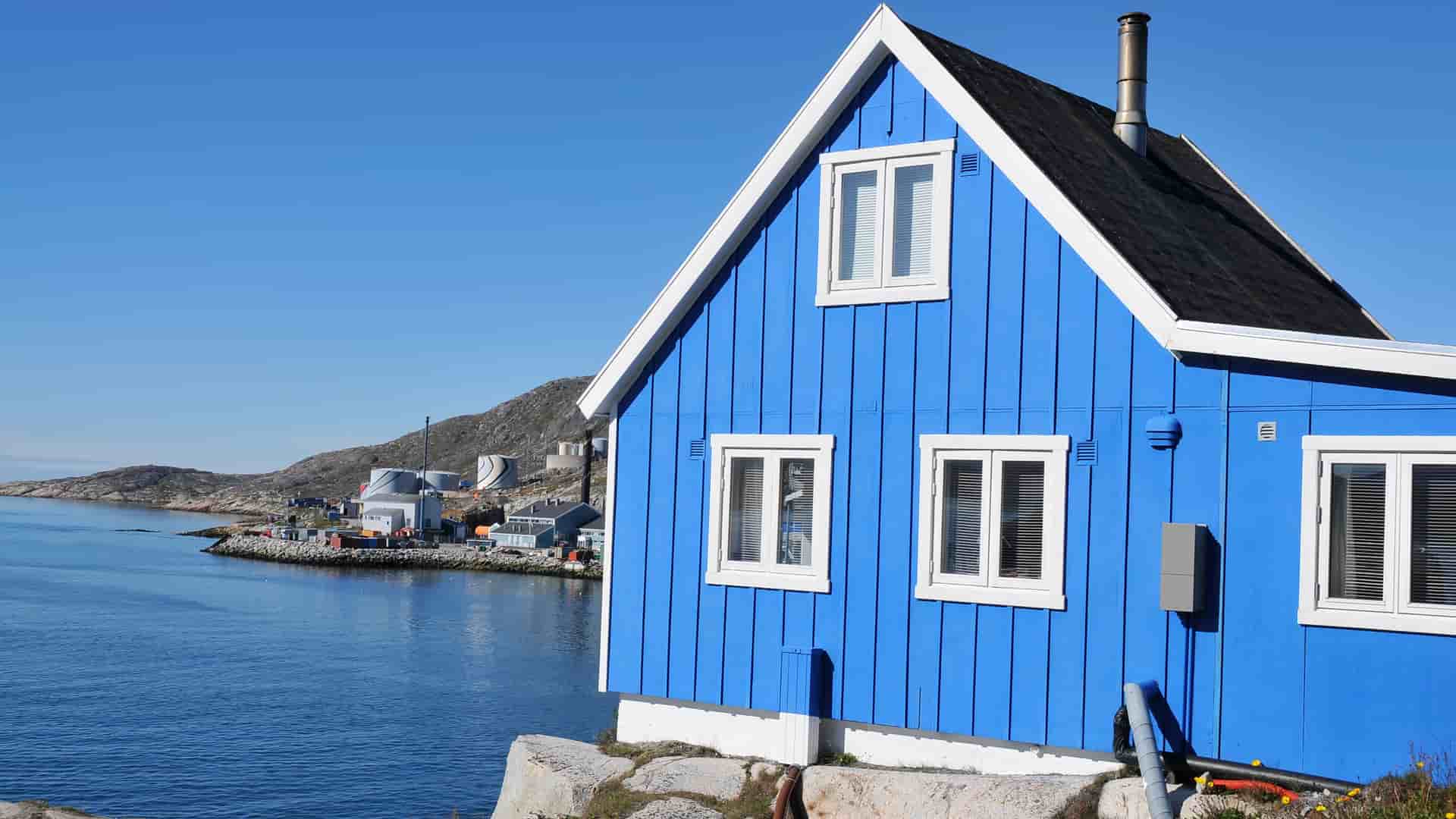 A striking blue wooden house with white trim overlooks the tranquil fjord and coastline of Qaqortoq, Greenland, a charming arctic town with colorful buildings.