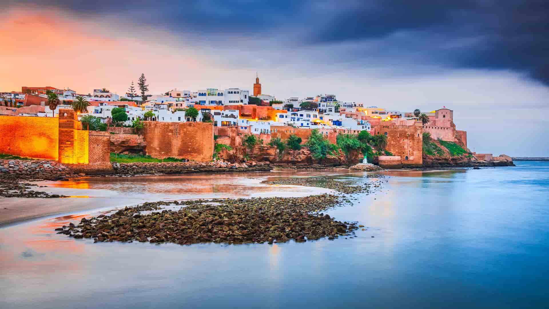 "A scenic view of the Kasbah of the Udayas in Rabat, Morocco, at dusk. The ancient, fortified walls and white-and-blue buildings are warmly lit, overlooking a calm body of water that reflects the vibrant orange, purple, and blue hues of the sunset sky.  "