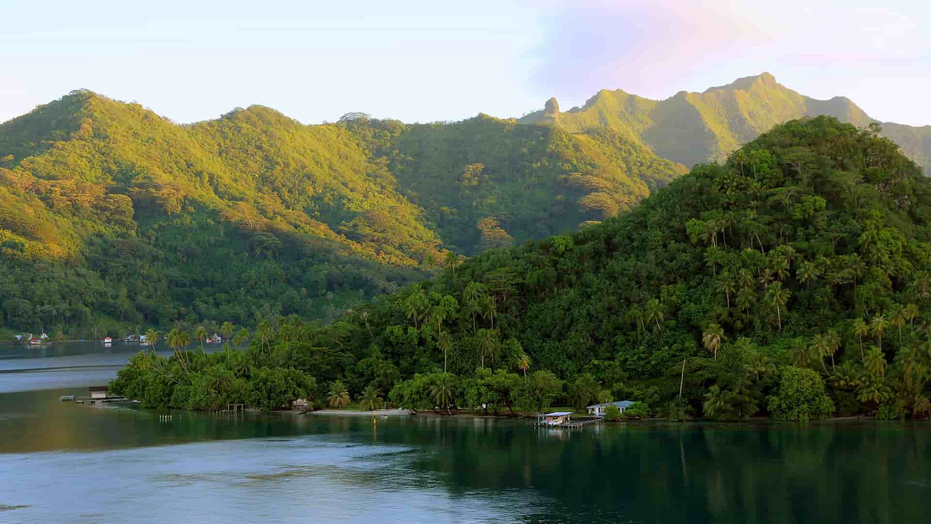 A breathtaking landscape of Raiatea Island, French Polynesia, showcasing lush, green volcanic mountains covered in tropical foliage descending into a tranquil turquoise lagoon.