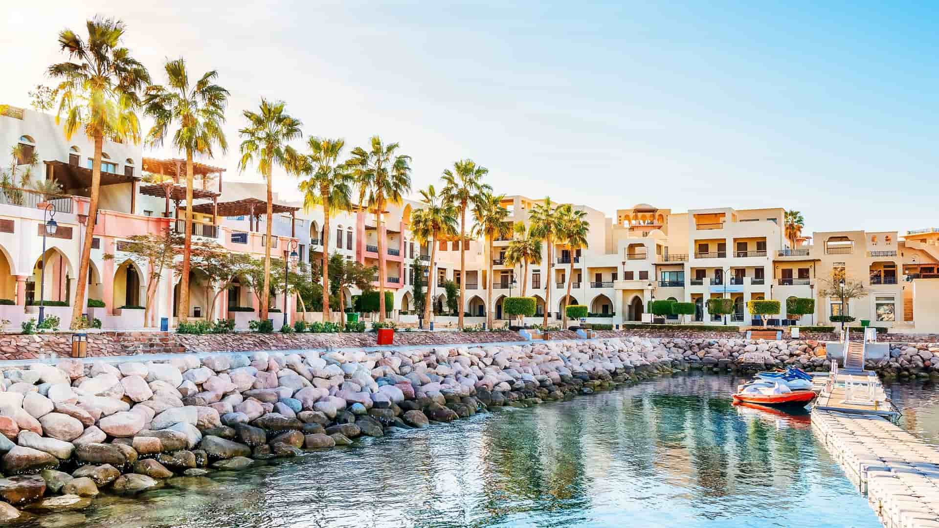 A beautiful view of the marina in Aqaba, Jordan, featuring traditional-style buildings and palm trees lining a stone-paved waterfront along the Red Sea.
