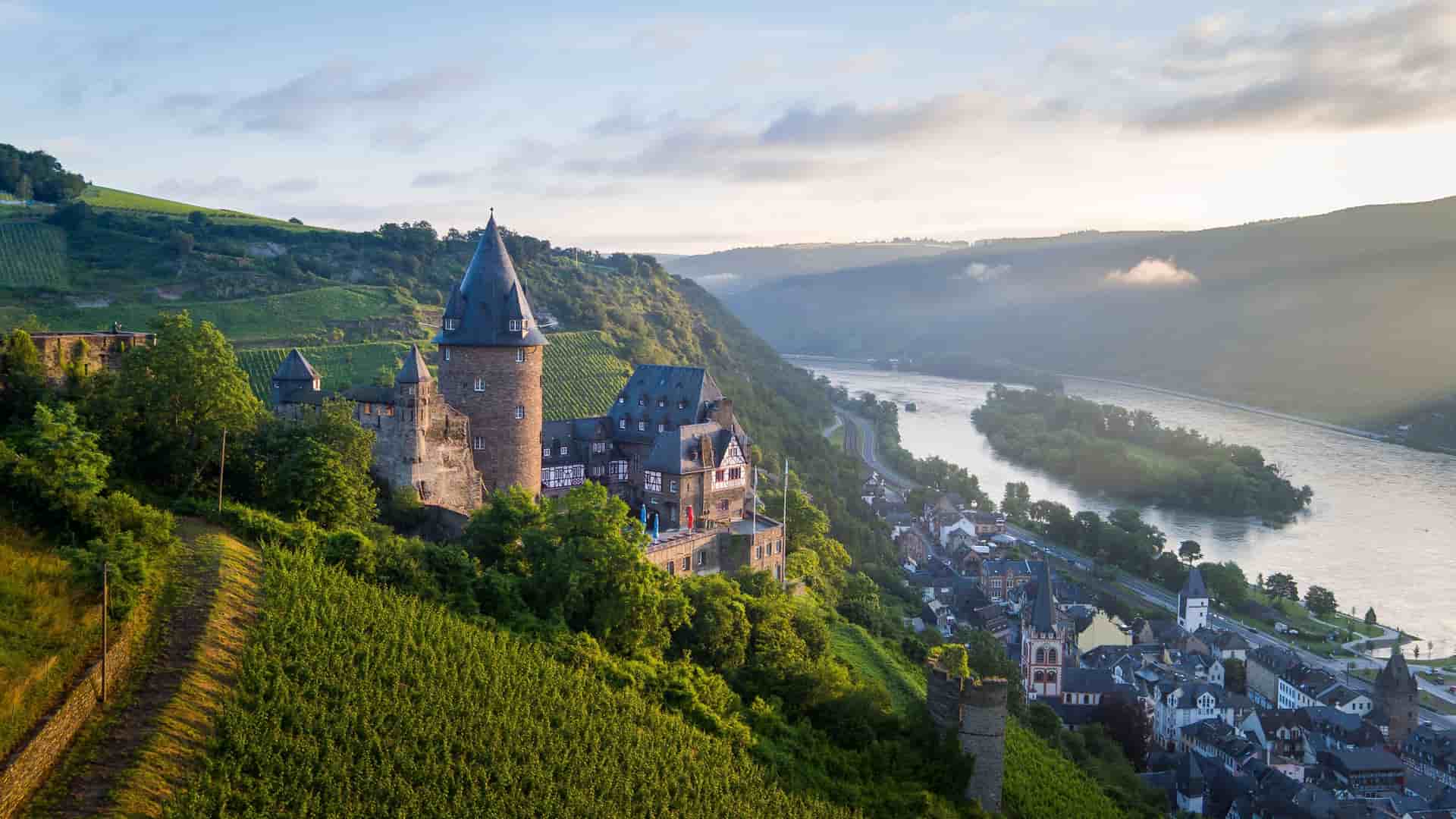 A scenic view of Stahleck Castle overlooking the town of Bacharach and the Rhine River in Germany, surrounded by vineyards and rolling green hills at sunrise.