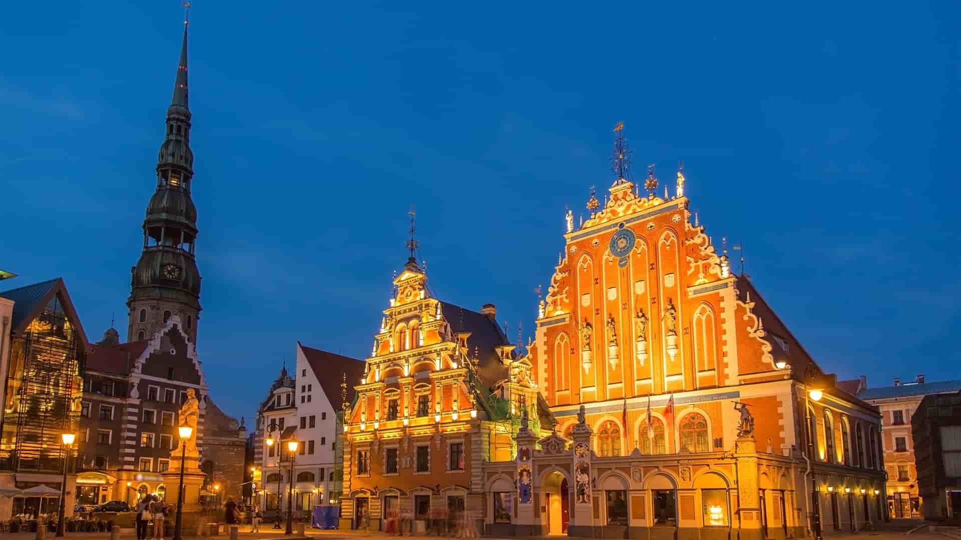 A nighttime shot of Riga's historic Old Town, featuring the beautifully illuminated House of the Blackheads and St. Peter's Church steeple against a deep blue sky.