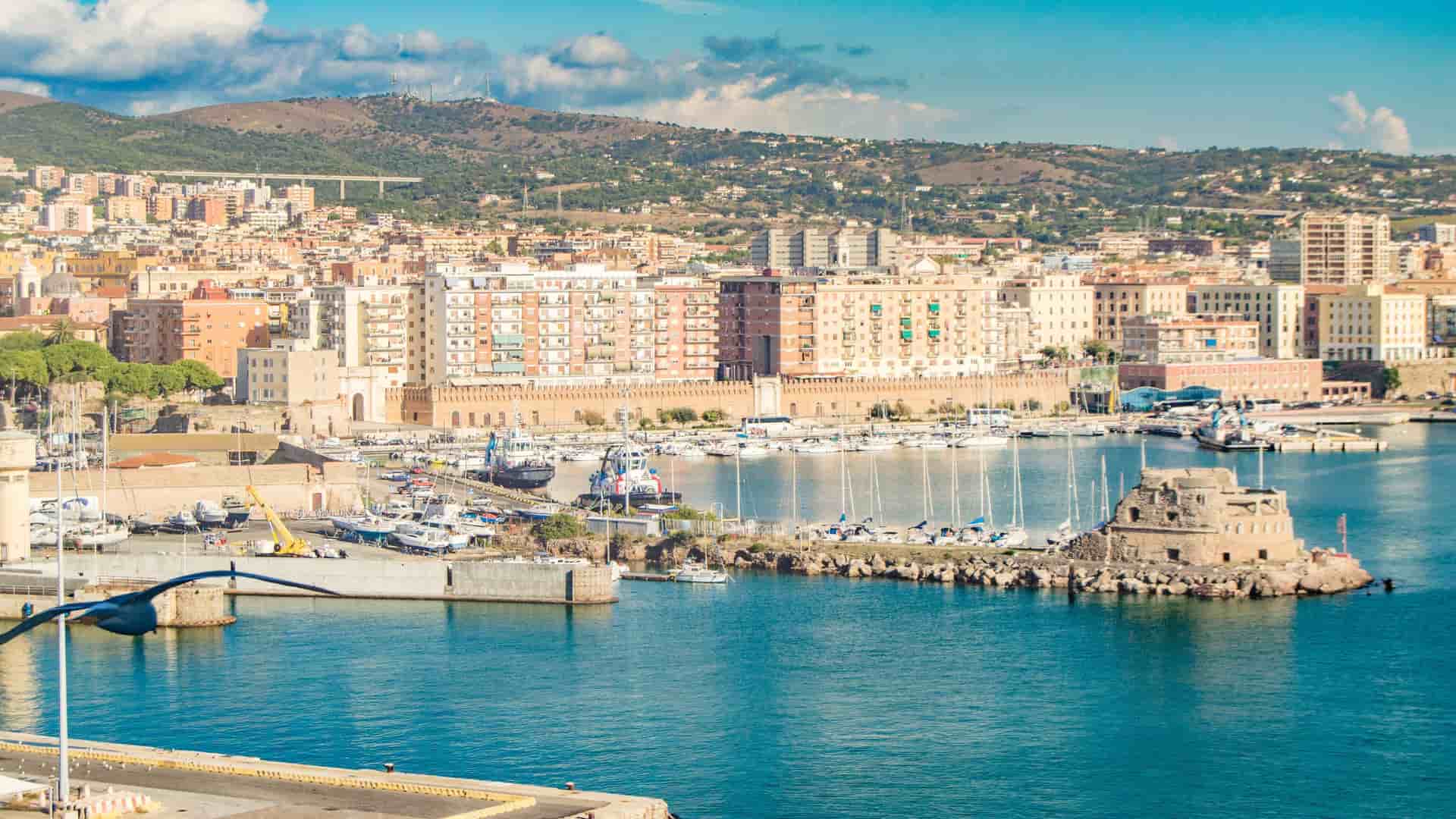 A beautiful view of the harbor and waterfront of Civitavecchia, Italy, with docked boats, historic buildings, and lush hills in the background.