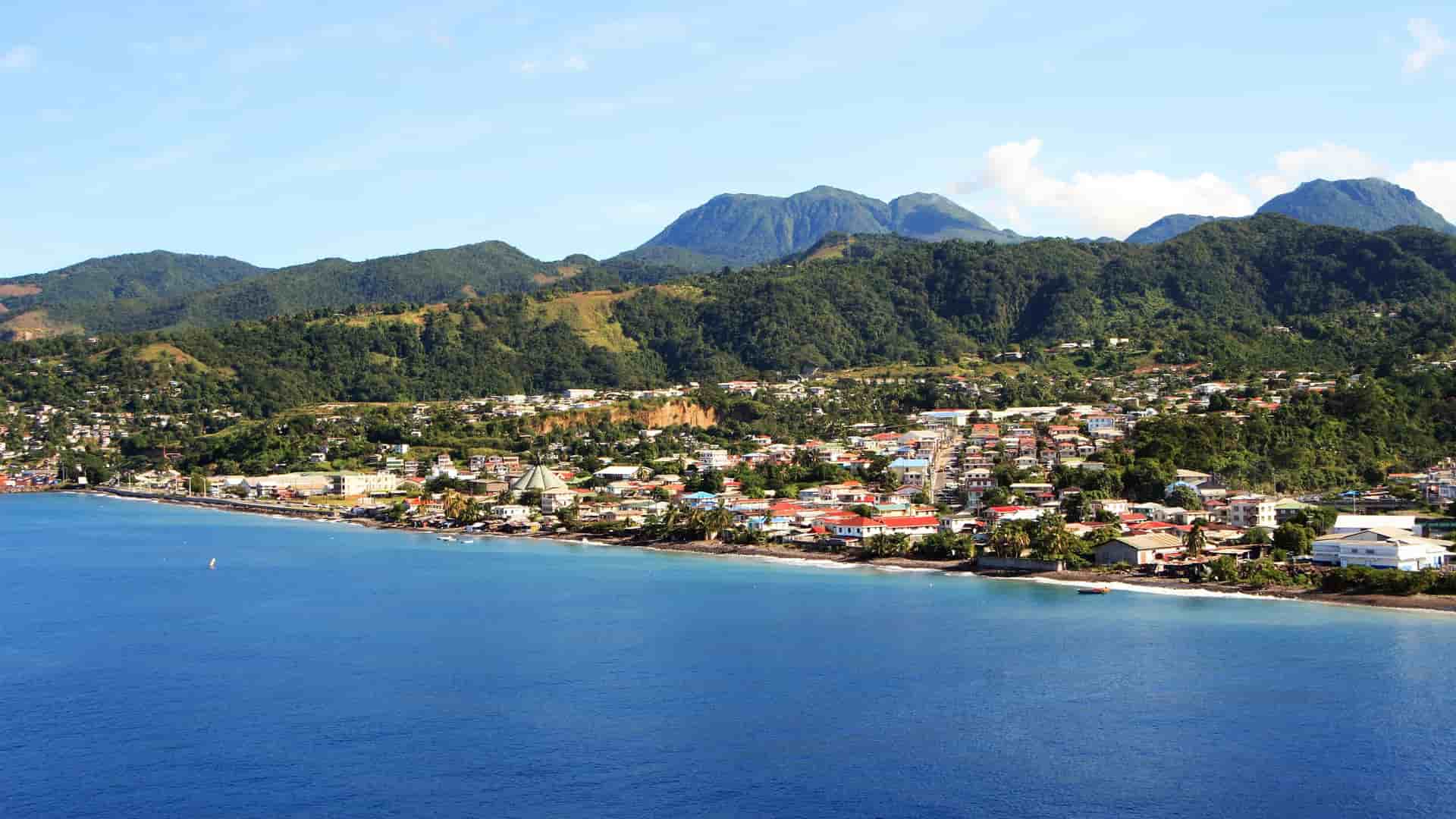 A beautiful panoramic view of Roseau, Dominica, with a colorful town nestled between the deep blue Caribbean Sea and a backdrop of lush green, volcanic mountains.