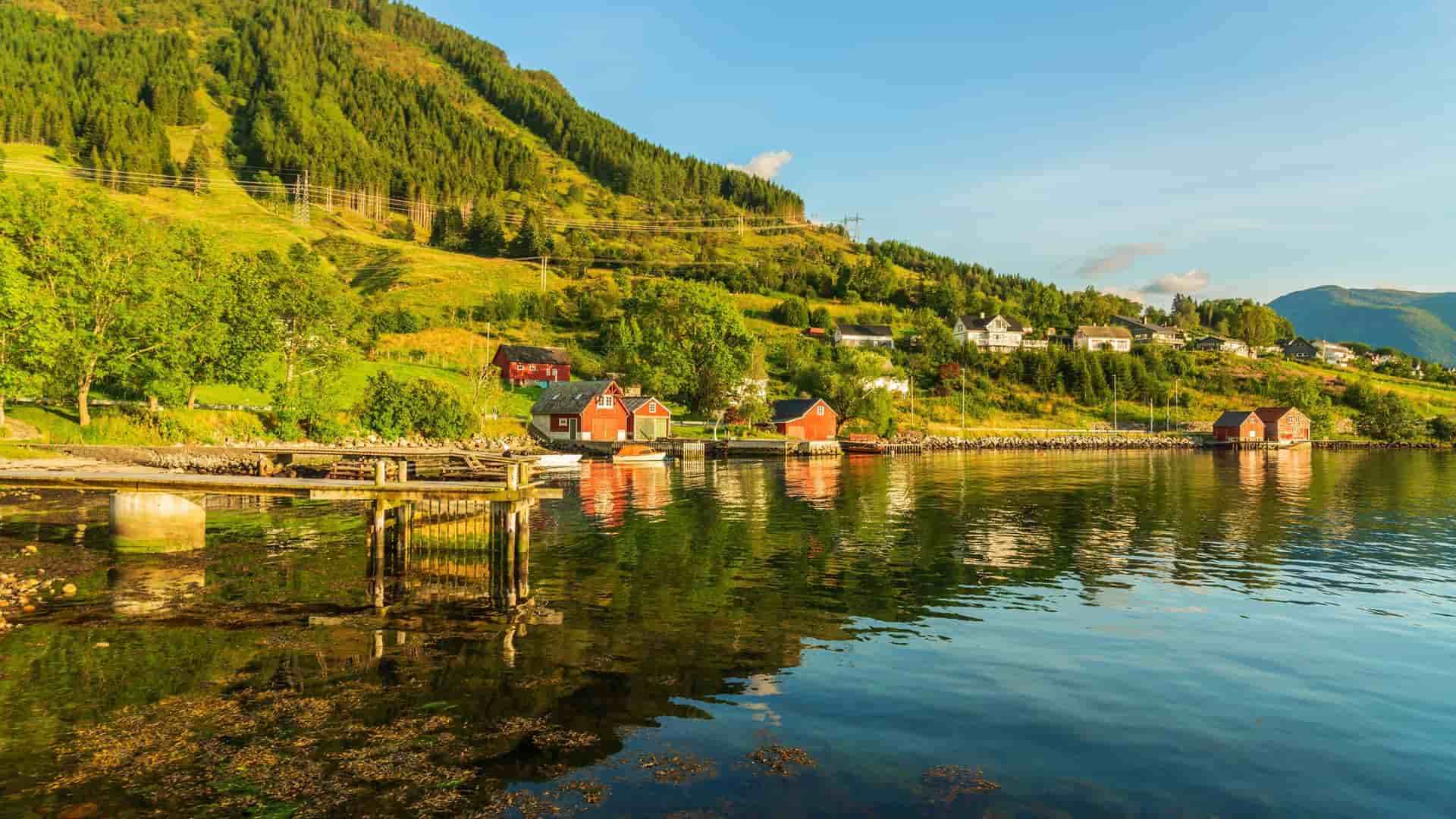 A serene view of Rosendal, Norway, with traditional red houses and a dock along the calm fjord, surrounded by lush green mountains under a bright sky.